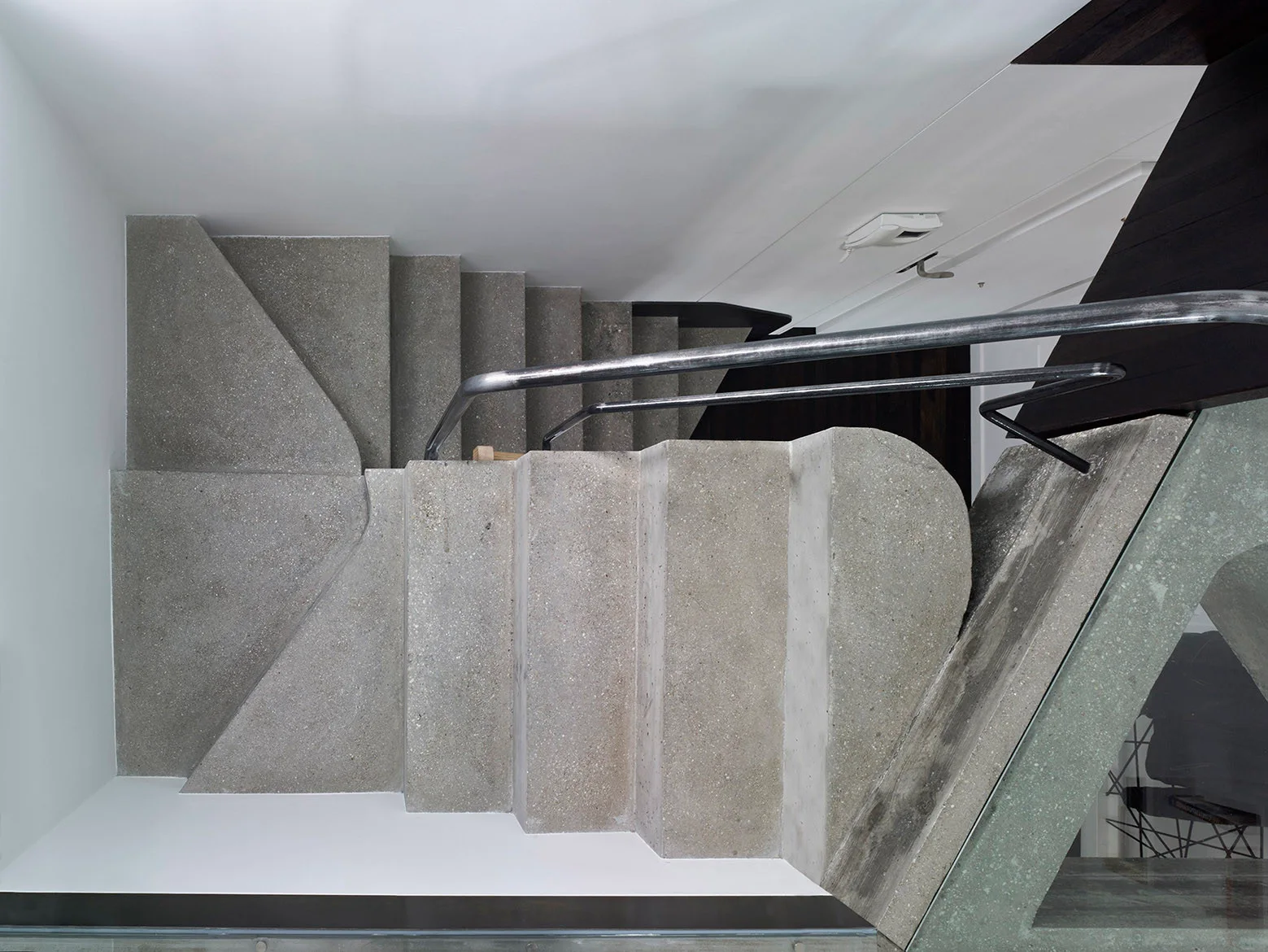 An overhead view of a geometric staircase with gray concrete steps and a curved metal handrail against plain white walls.