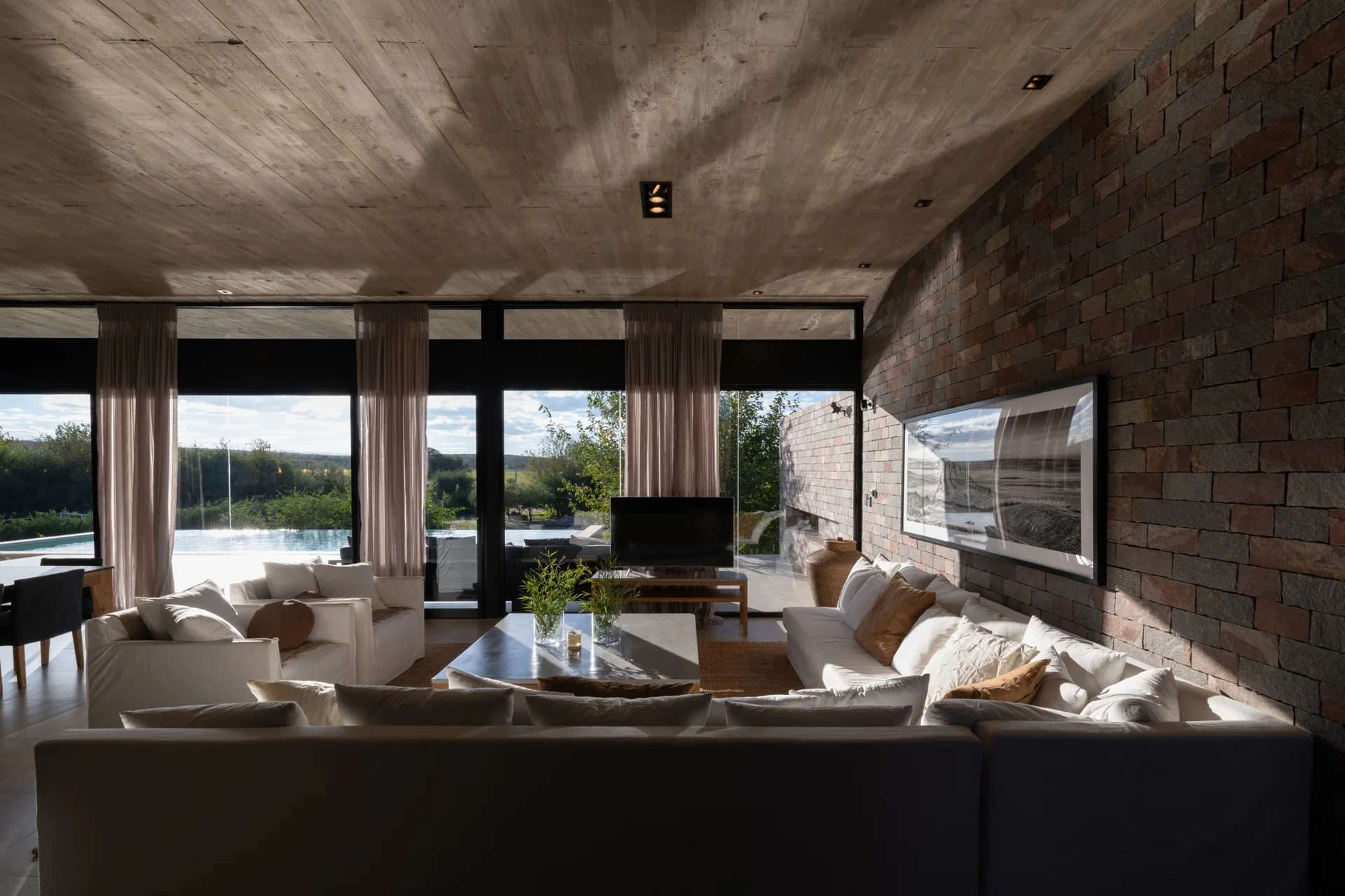 White sectional sofas face a large glass wall in a living room featuring an exposed concrete ceiling and a textured stone accent wall.
