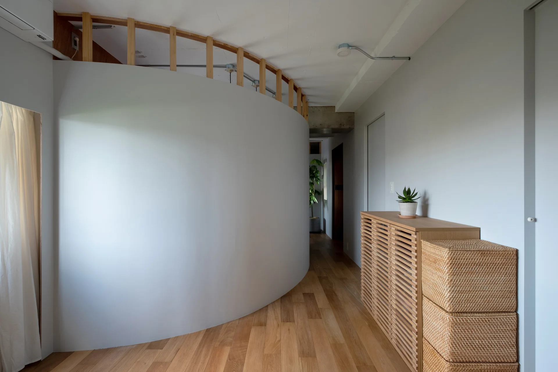A minimalist hallway features a curved white partition wall, a slatted wooden console, and light hardwood floors.