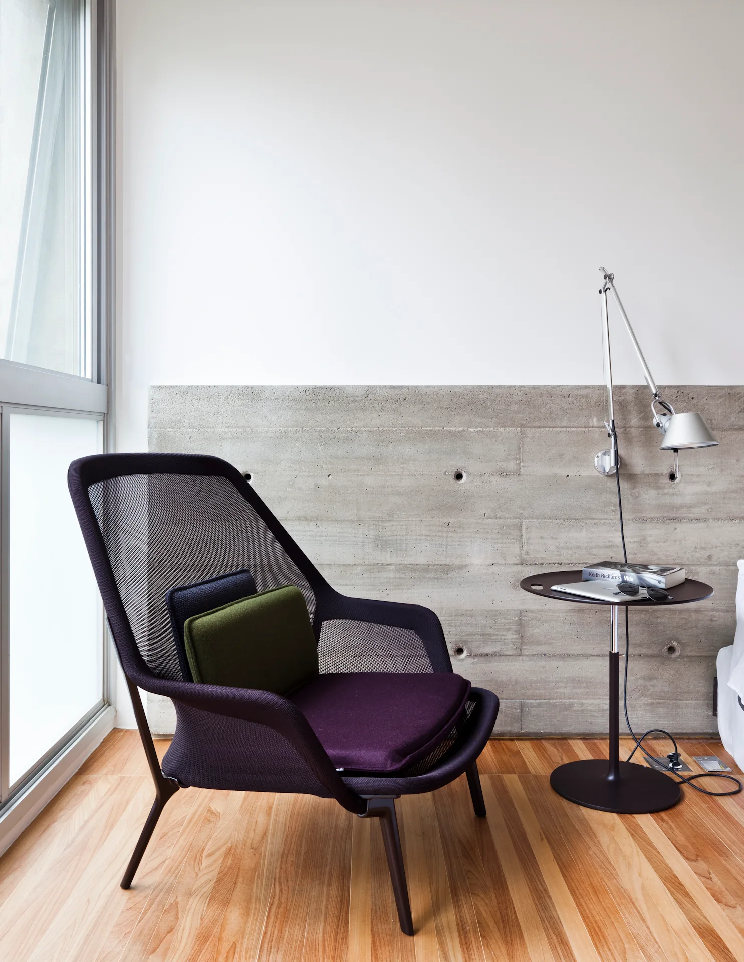 A modern mesh lounge chair with purple and green cushions sits next to a black pedestal table and a wall lamp against a concrete wall.