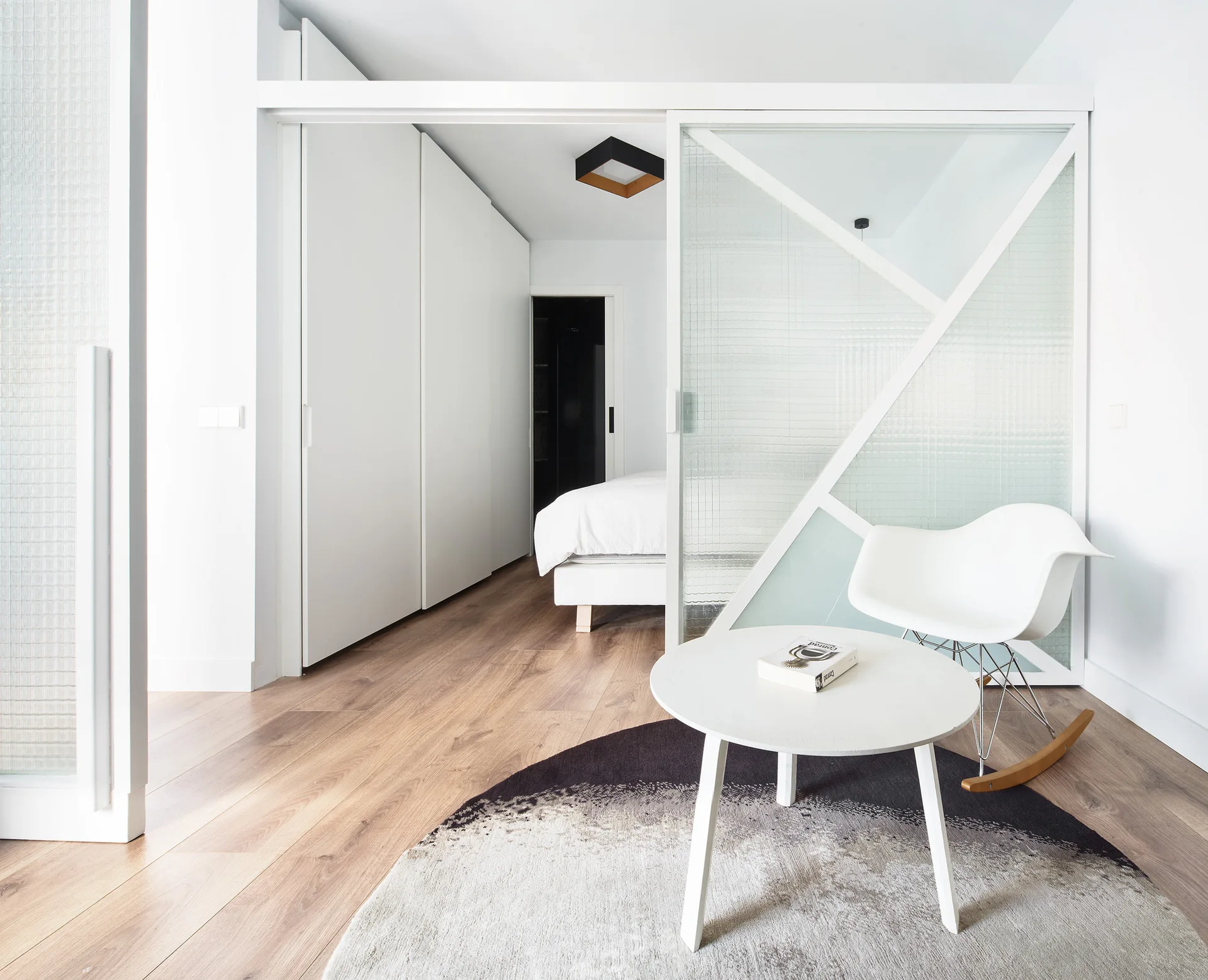 A modern bedroom featuring a geometric sliding glass partition next to a white rocking chair, round coffee table, and wood flooring.