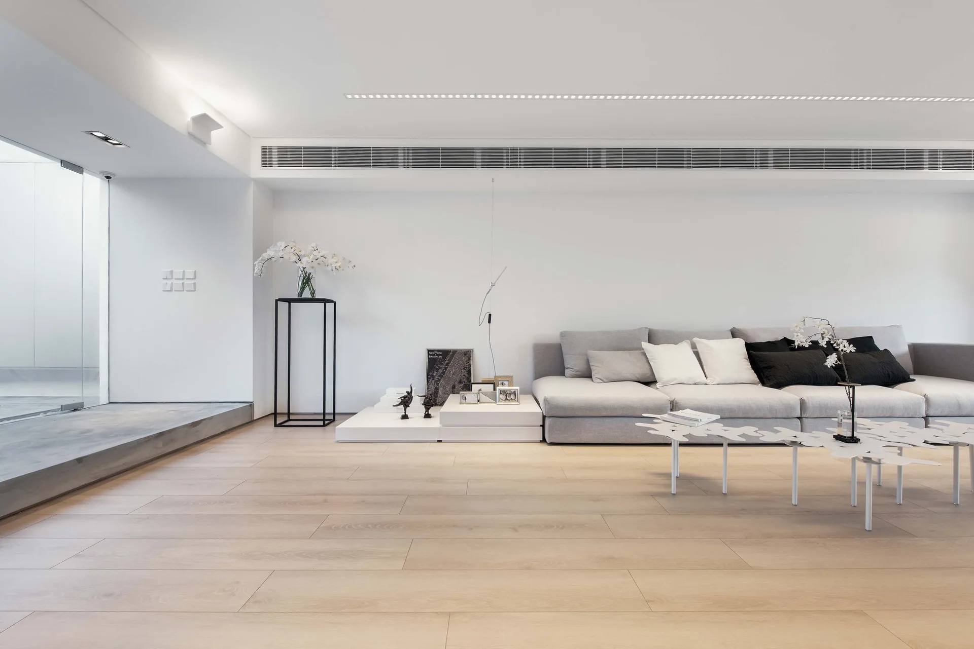 A minimalist living room with a large grey sectional sofa, low white display platforms, sculptural white coffee tables, and light wood floors.