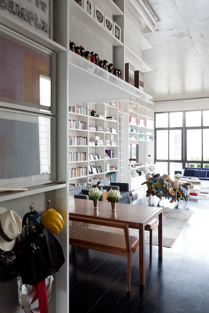 An open loft features a wooden dining table, a cane bench, and a towering white built-in bookcase with an integrated library ladder.