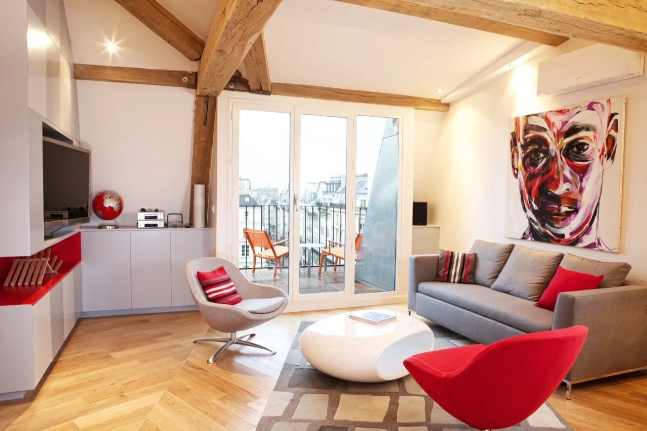 A contemporary living room with exposed wood ceiling beams, a grey sofa, a glossy white coffee table, and red accent chairs.