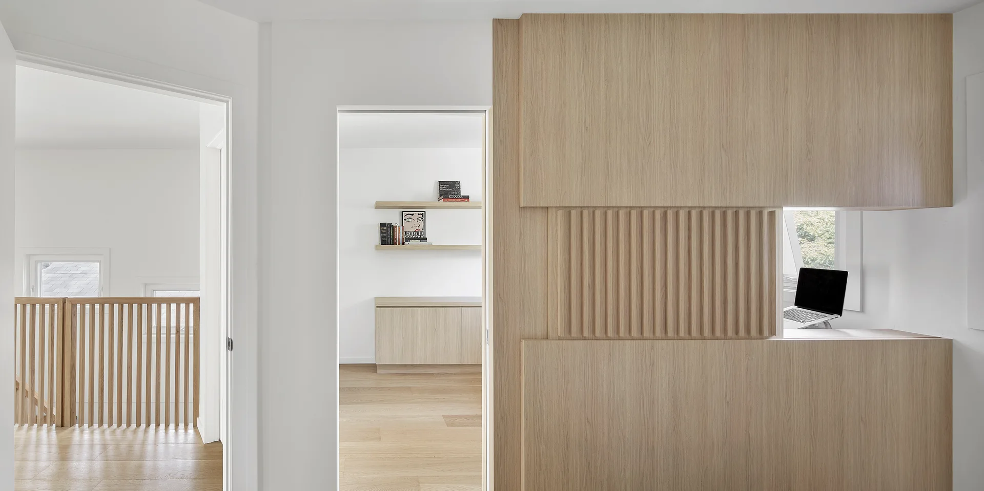 A minimalist hallway features a light wood partition wall with fluted paneling, a small cutout window, and open doorways showing adjacent rooms.