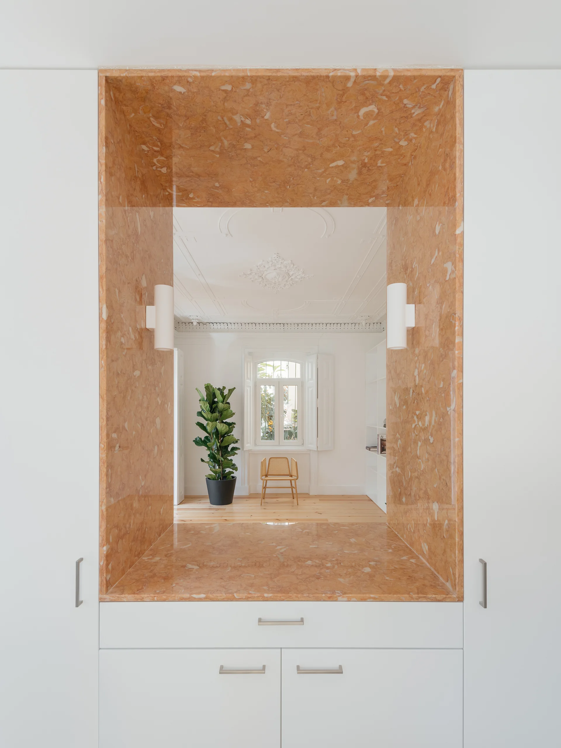 A view through an orange veined marble niche in white built-in cabinets looking into a bright room with a cane chair and potted plant.