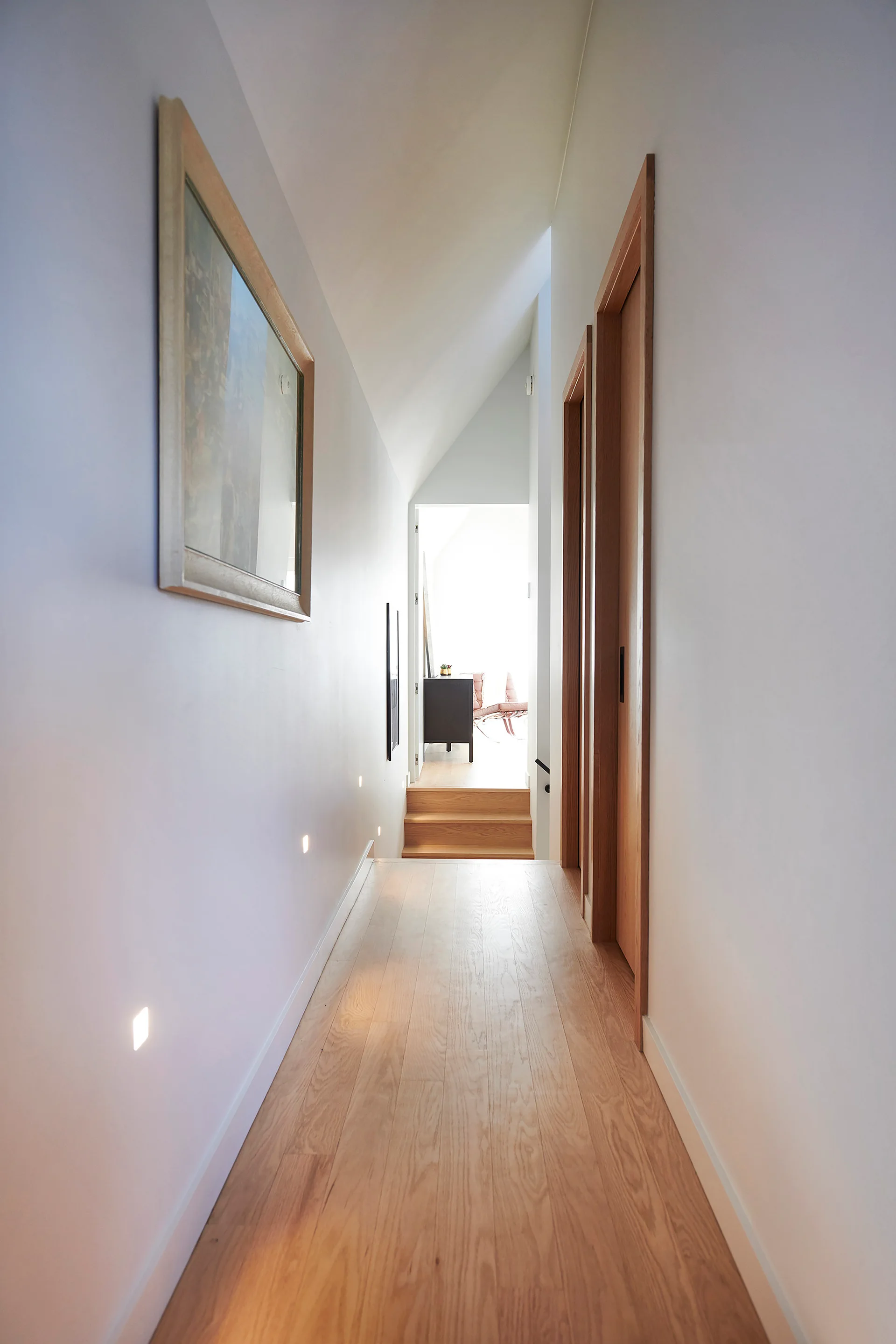 A narrow minimalist hallway features light wood floors, matching wood doors, a sloped white ceiling, and low-level square recessed wall lights.