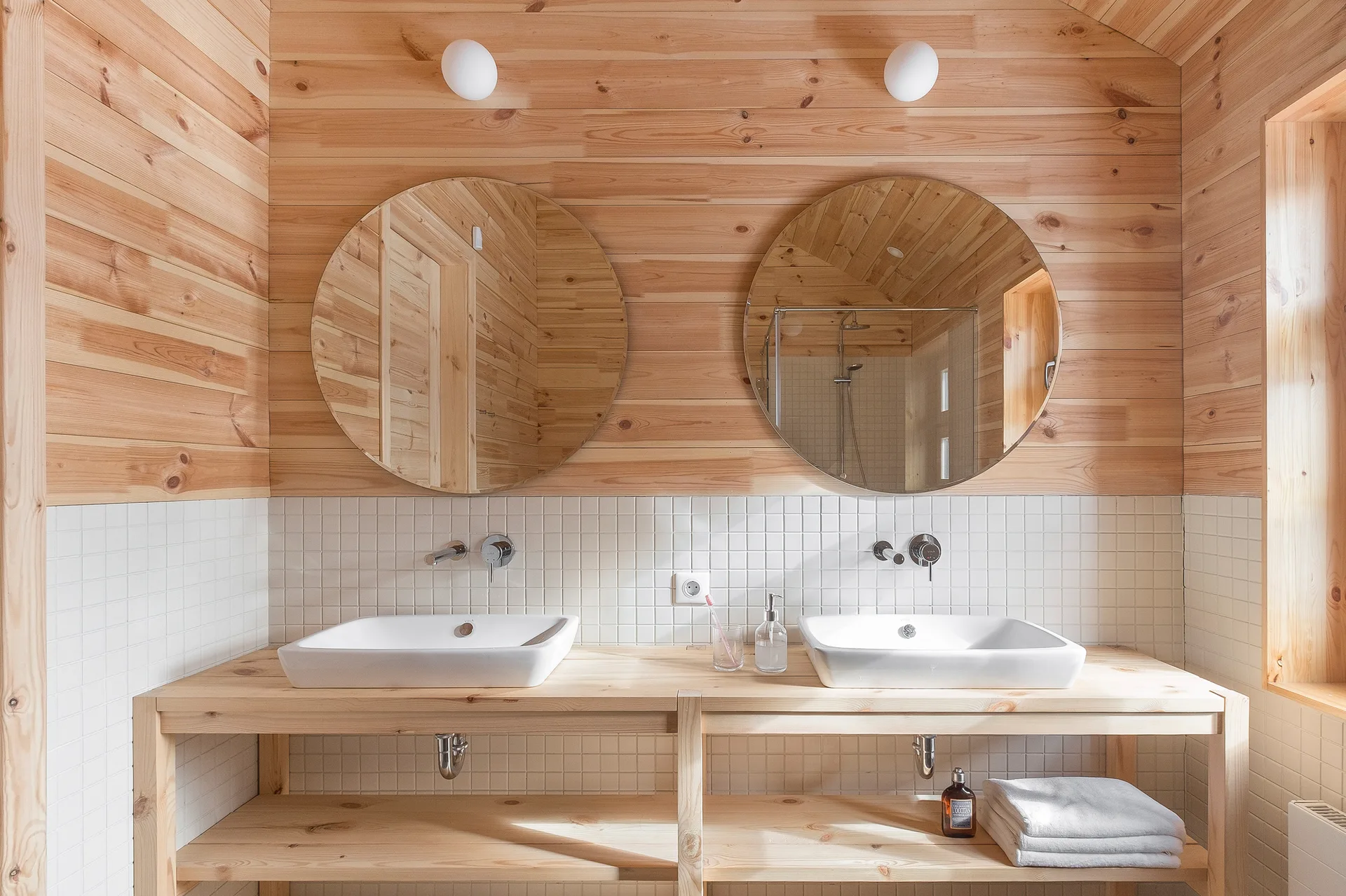 A bathroom features a double wood vanity with white vessel sinks below two round mirrors, set against white grid tile and wood-paneled walls.