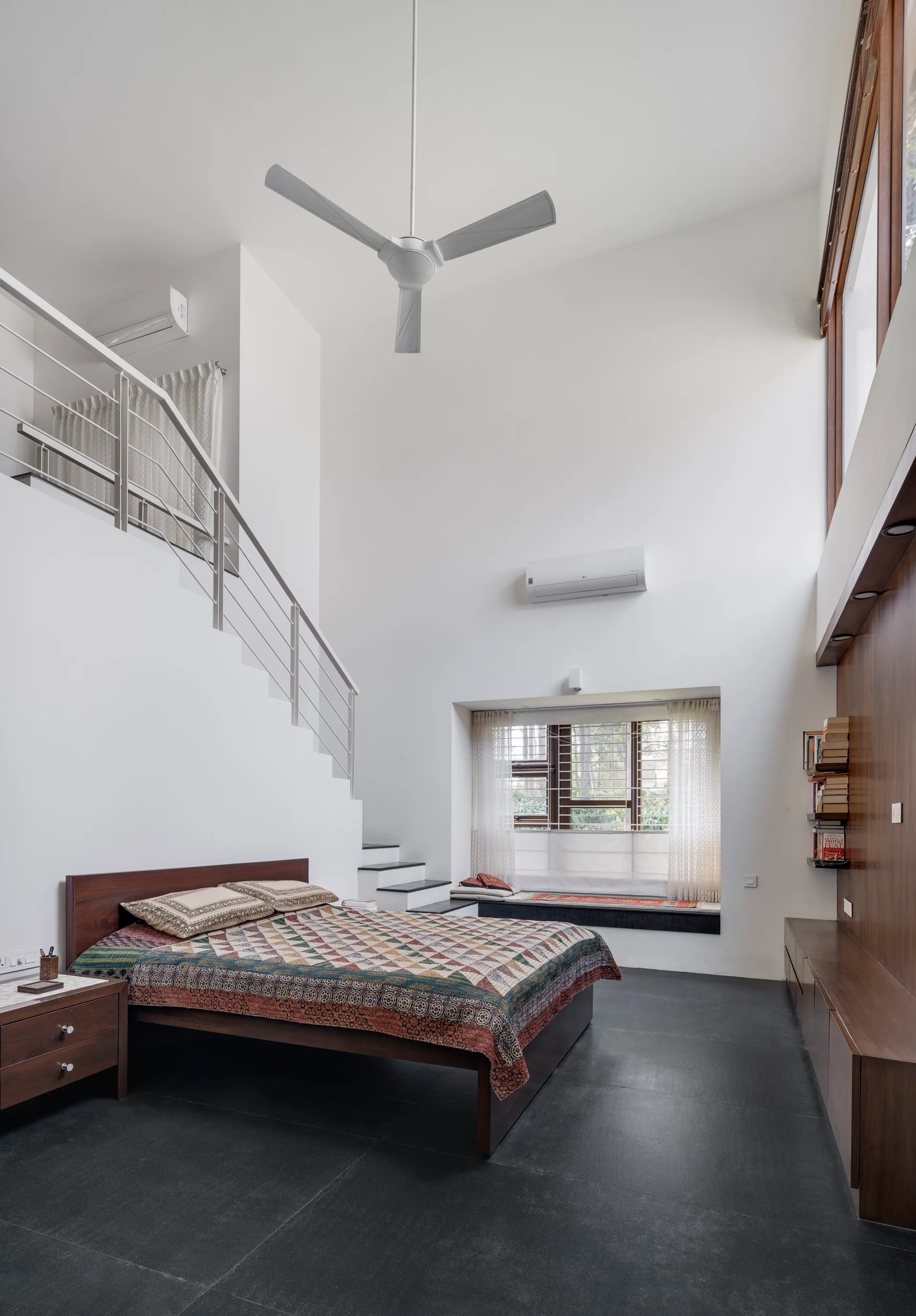 A high-ceilinged minimalist bedroom featuring a wooden platform bed, an integrated staircase with metal railing, and a dark wood accent wall.