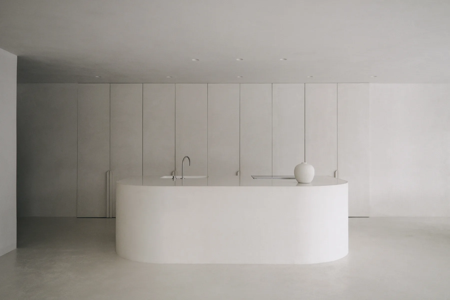 A minimalist monochromatic kitchen features a curved off-white island in front of a seamless wall of floor-to-ceiling cabinetry.