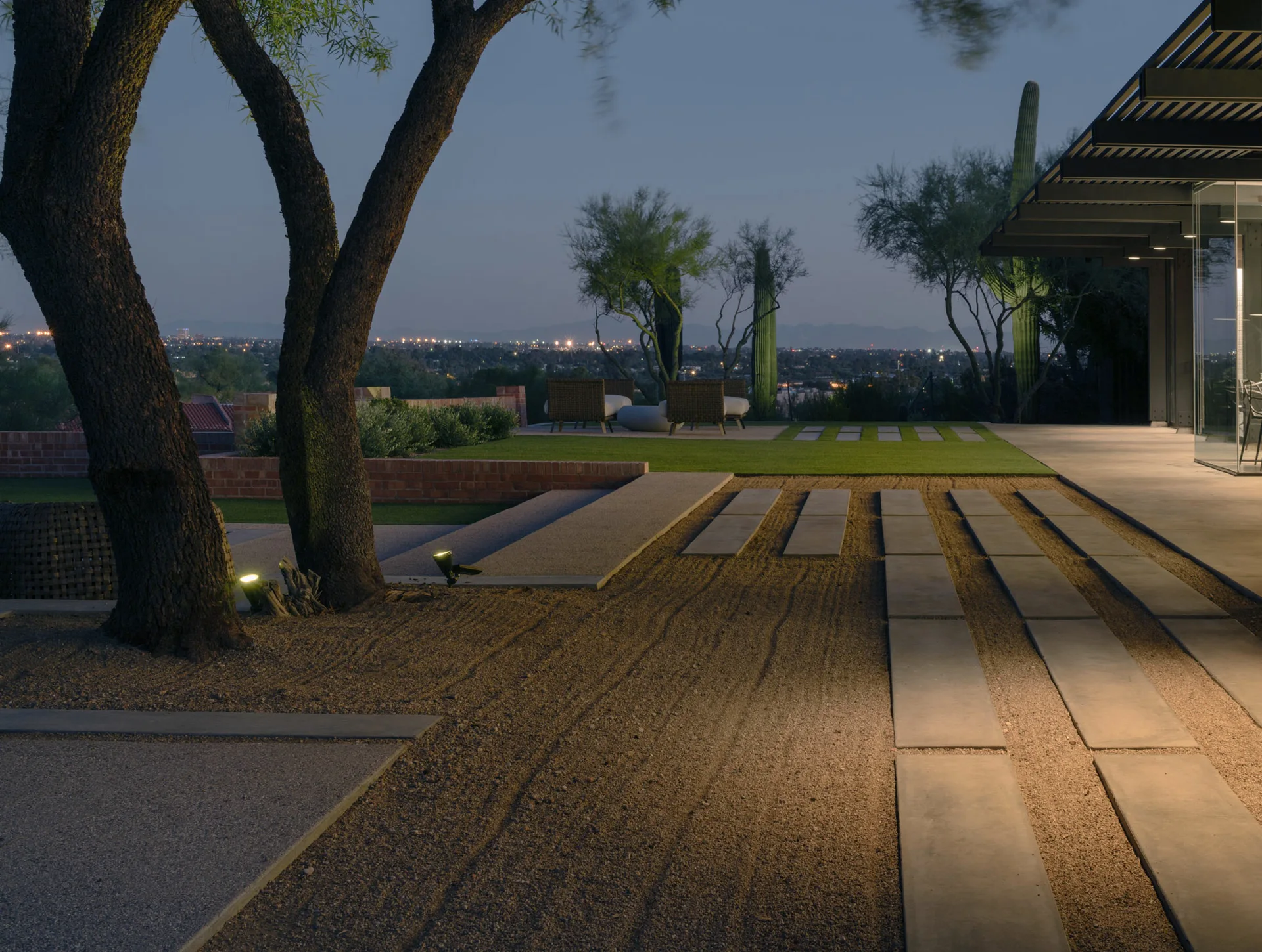 A twilight view of a desert garden featuring long concrete stepping stones set in gravel, a brick retaining wall, and distant saguaro cacti.