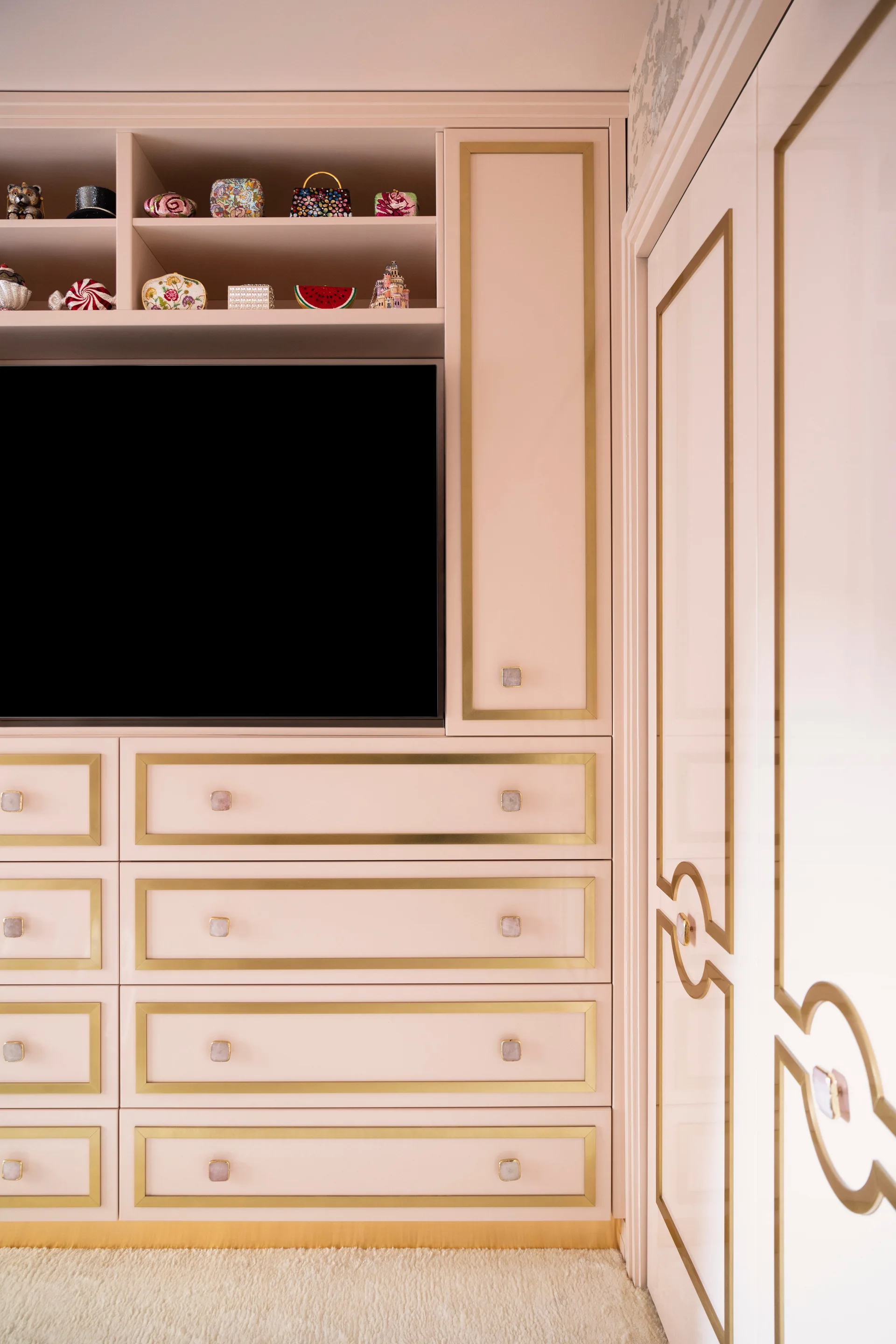 A detail of blush pink built-in cabinetry featuring brass trim on the drawers, square stone knobs, an integrated television, and open top shelves.