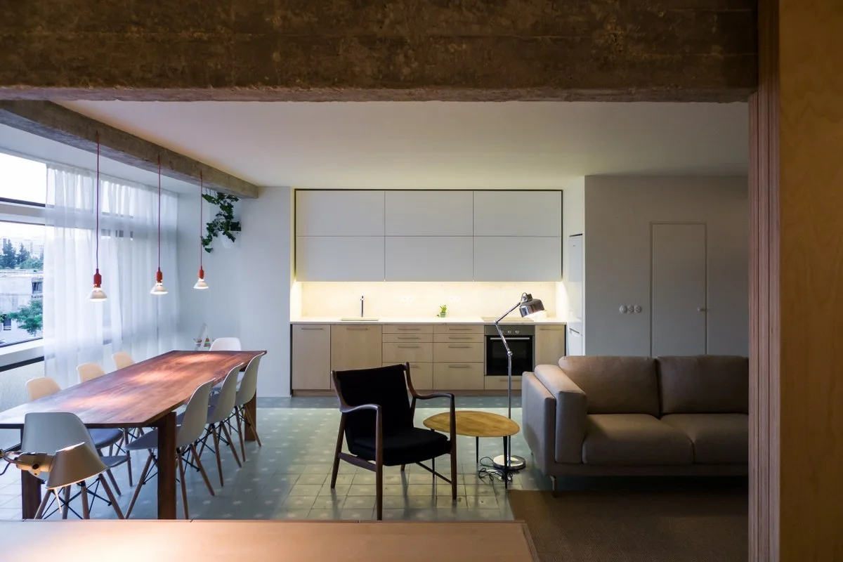 A minimalist open-plan apartment interior shows a wood dining table under red pendant lights, light wood kitchen cabinets, and blue-grey tiled floors.