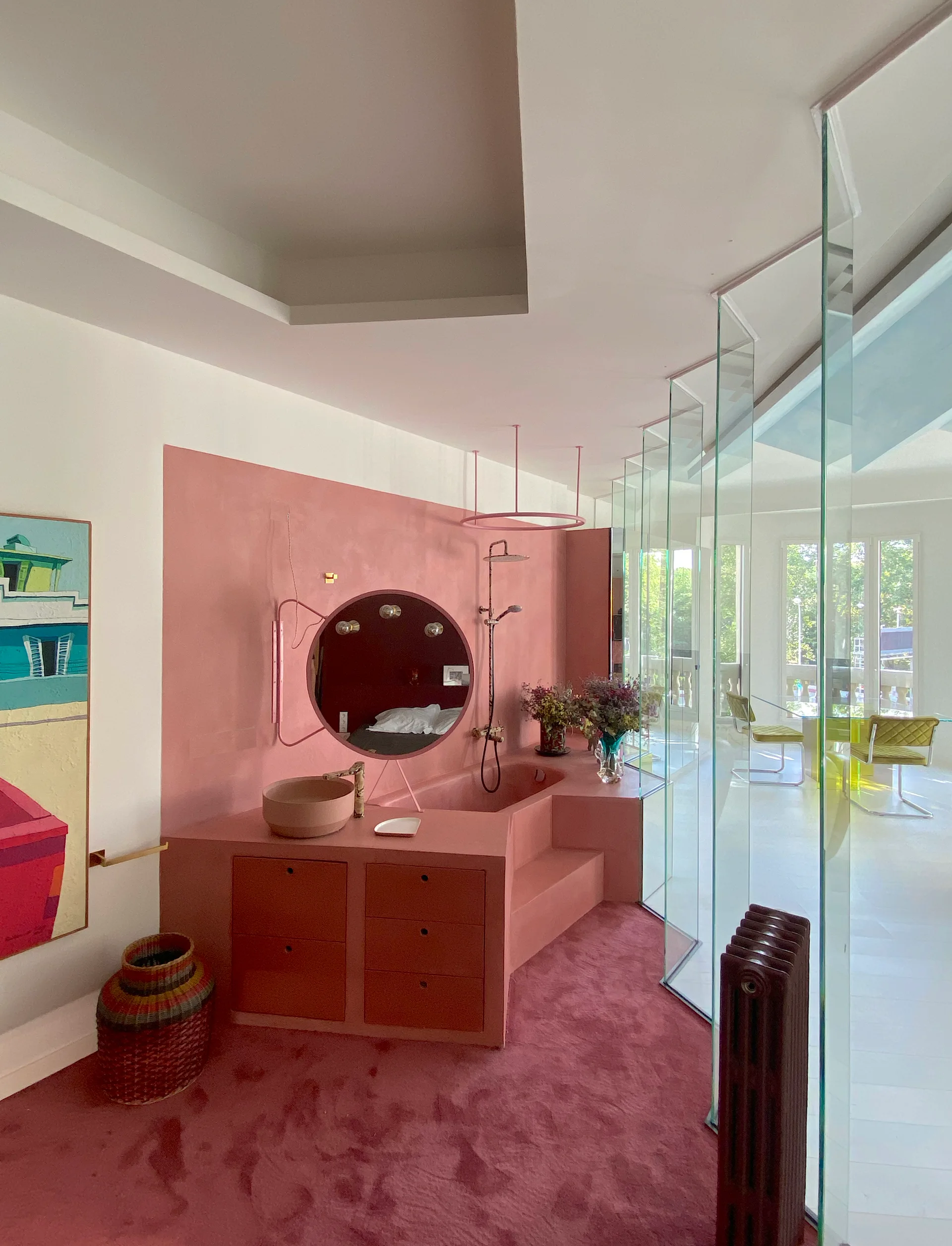 A monochromatic pink bathroom suite features a built-in vanity and integrated bathtub separated from a dining area by vertical glass panels.