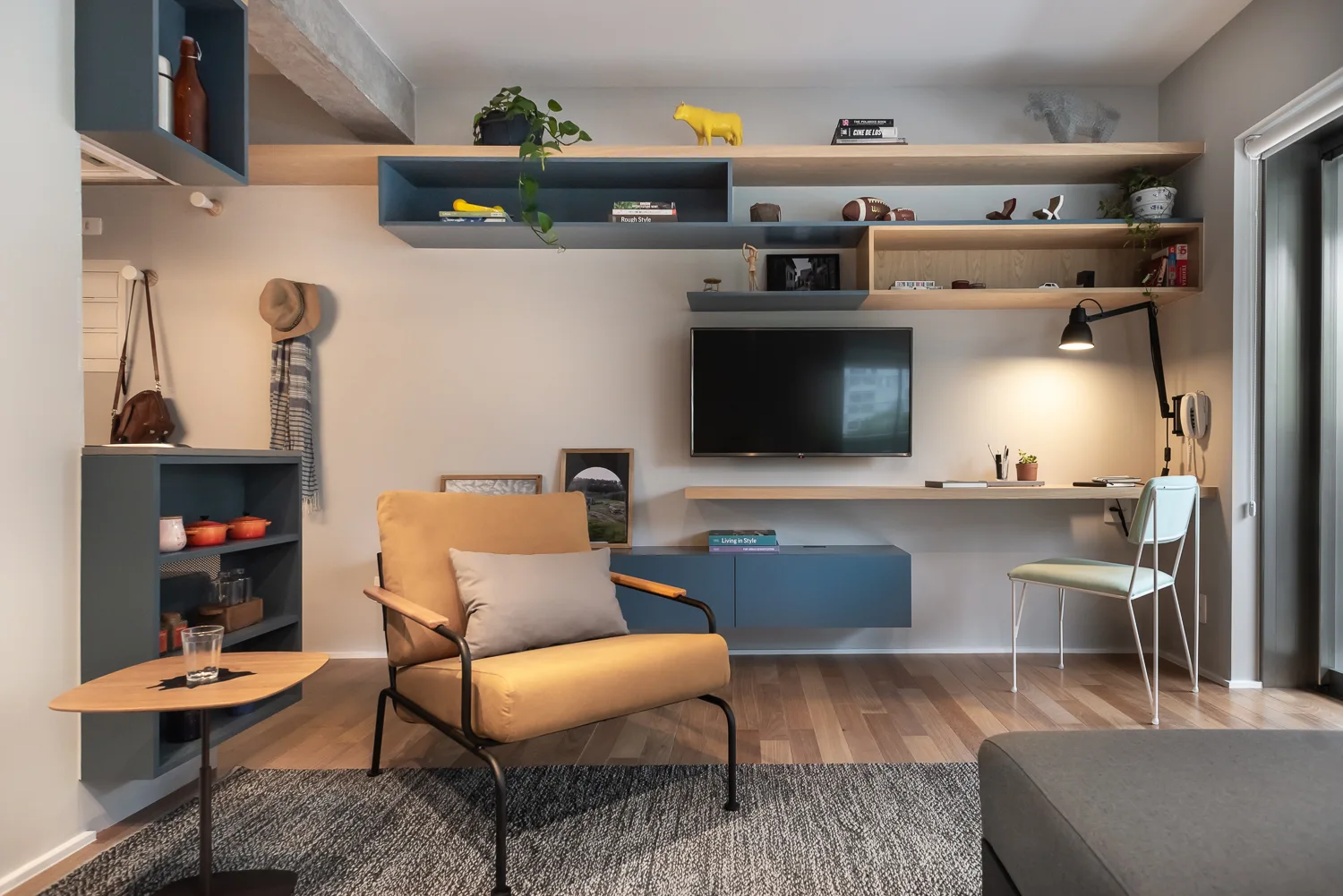 A contemporary living room featuring a yellow armchair on a textured rug next to a custom blue and wood floating shelving unit with an integrated desk.