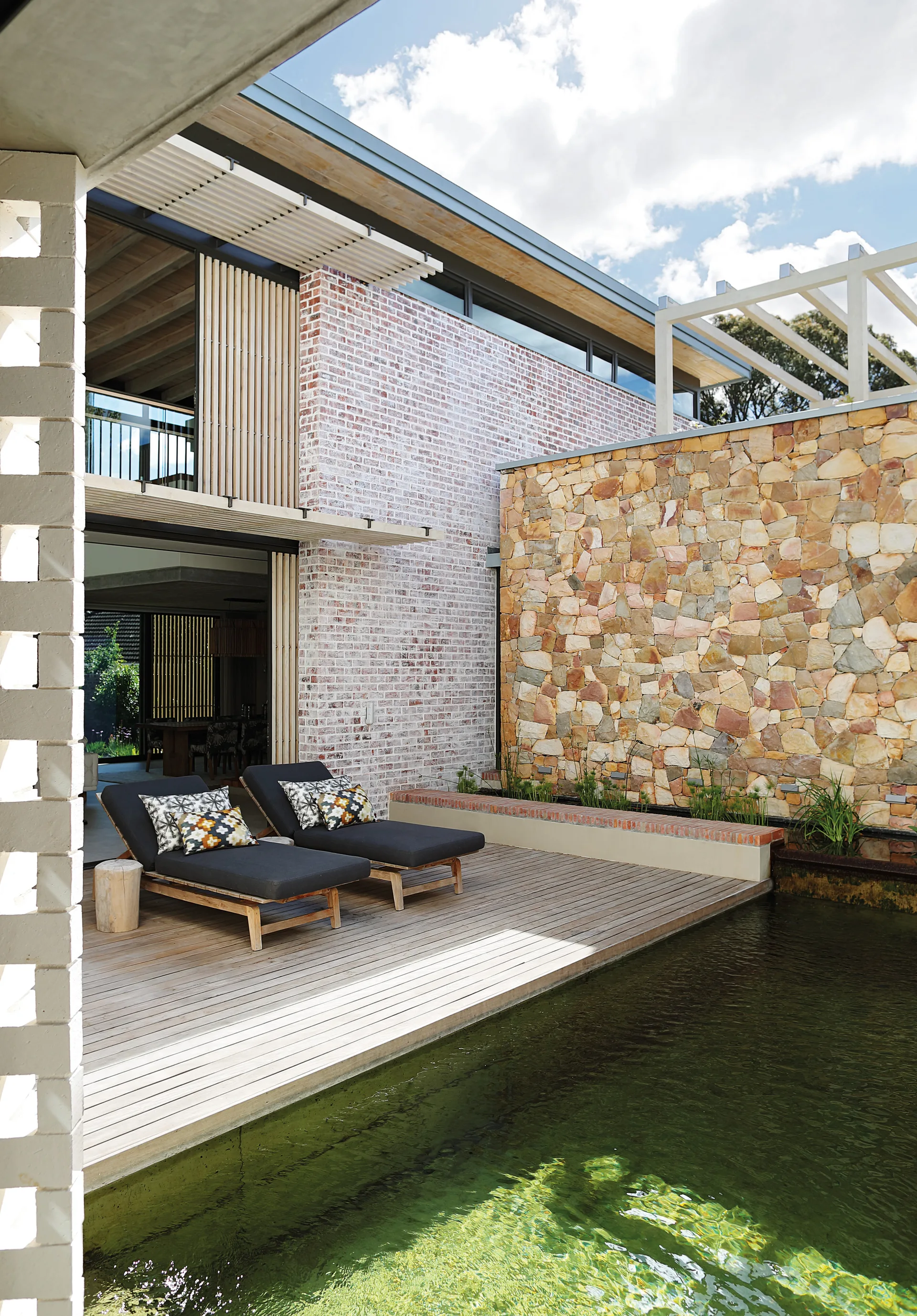 Two charcoal lounge chairs sit on a light wood deck beside a swimming pool, bordered by a whitewashed brick exterior and a stacked stone wall.