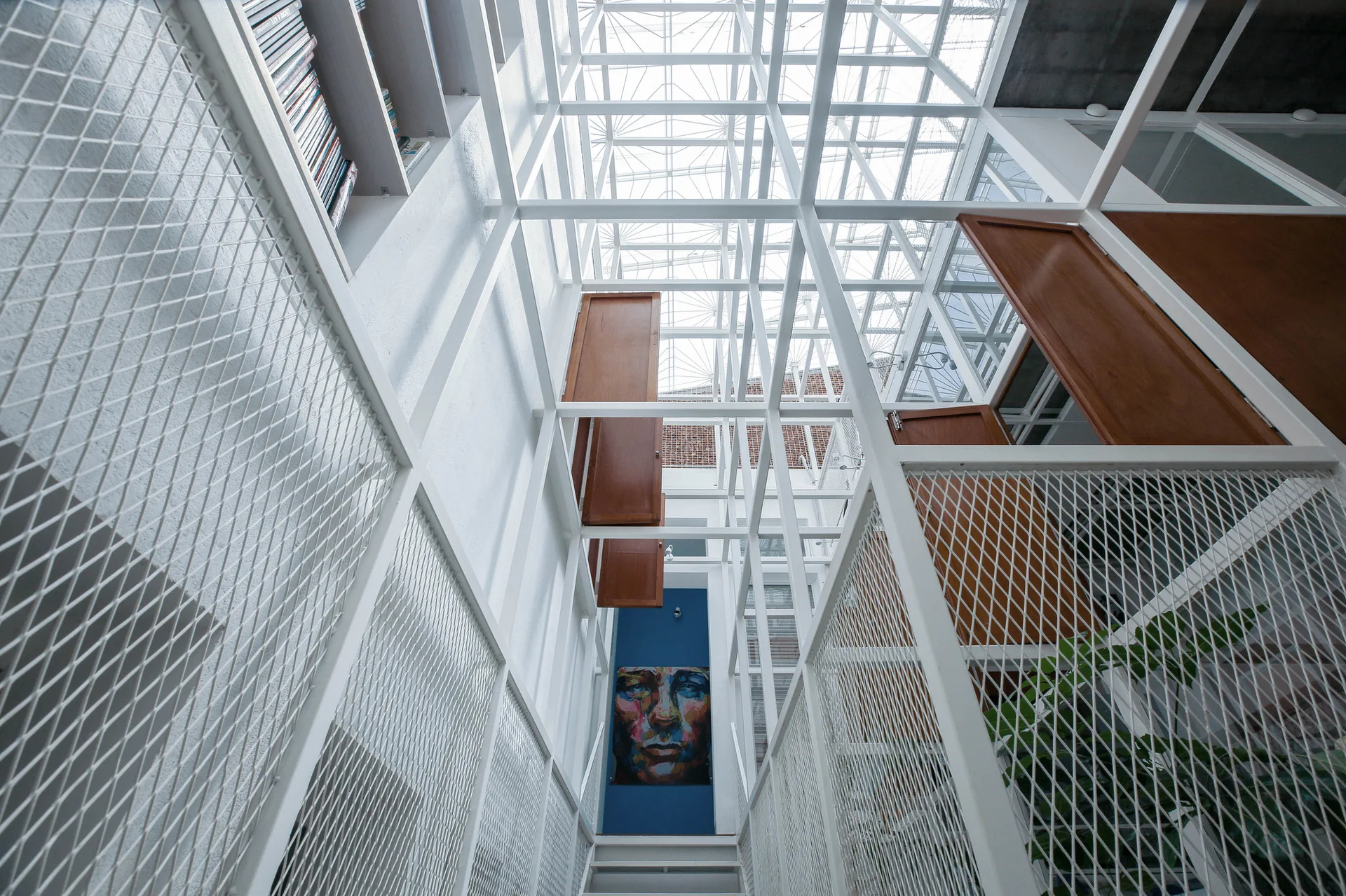 A vertical hallway featuring a white metal grid framework, wire mesh railings, a glass skylight ceiling, and several wooden shutter doors.