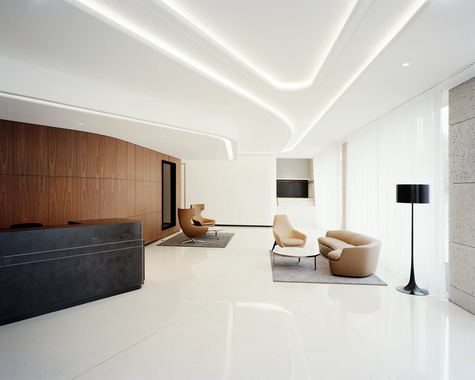 A minimalist reception lobby features a dark stone reception desk, wood-paneled walls, tan seating areas, and curved cove lighting in a white ceiling.