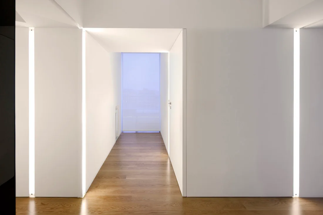 A minimalist hallway featuring white walls, warm wood flooring, and vertical recessed LED light strips embedded in the plaster surfaces.