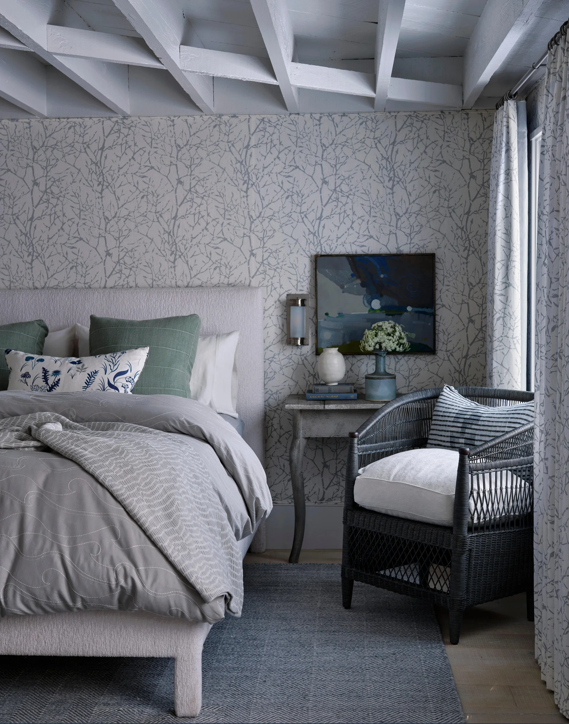 A bedroom featuring an upholstered bed, a dark wicker armchair, and delicate branch-patterned wallpaper beneath white exposed ceiling beams.