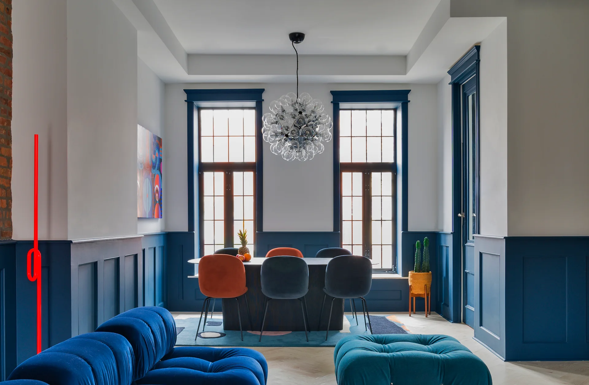 A dining room features deep blue wainscoting and matching window trim, modern colorful chairs around an oval table, and a glass bubble chandelier.