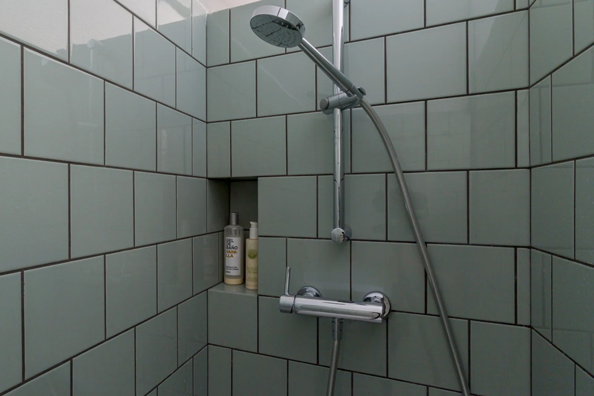 A close-up of a shower enclosure featuring glossy sage green wall tiles, dark grout lines, chrome fixtures, and a recessed storage niche.