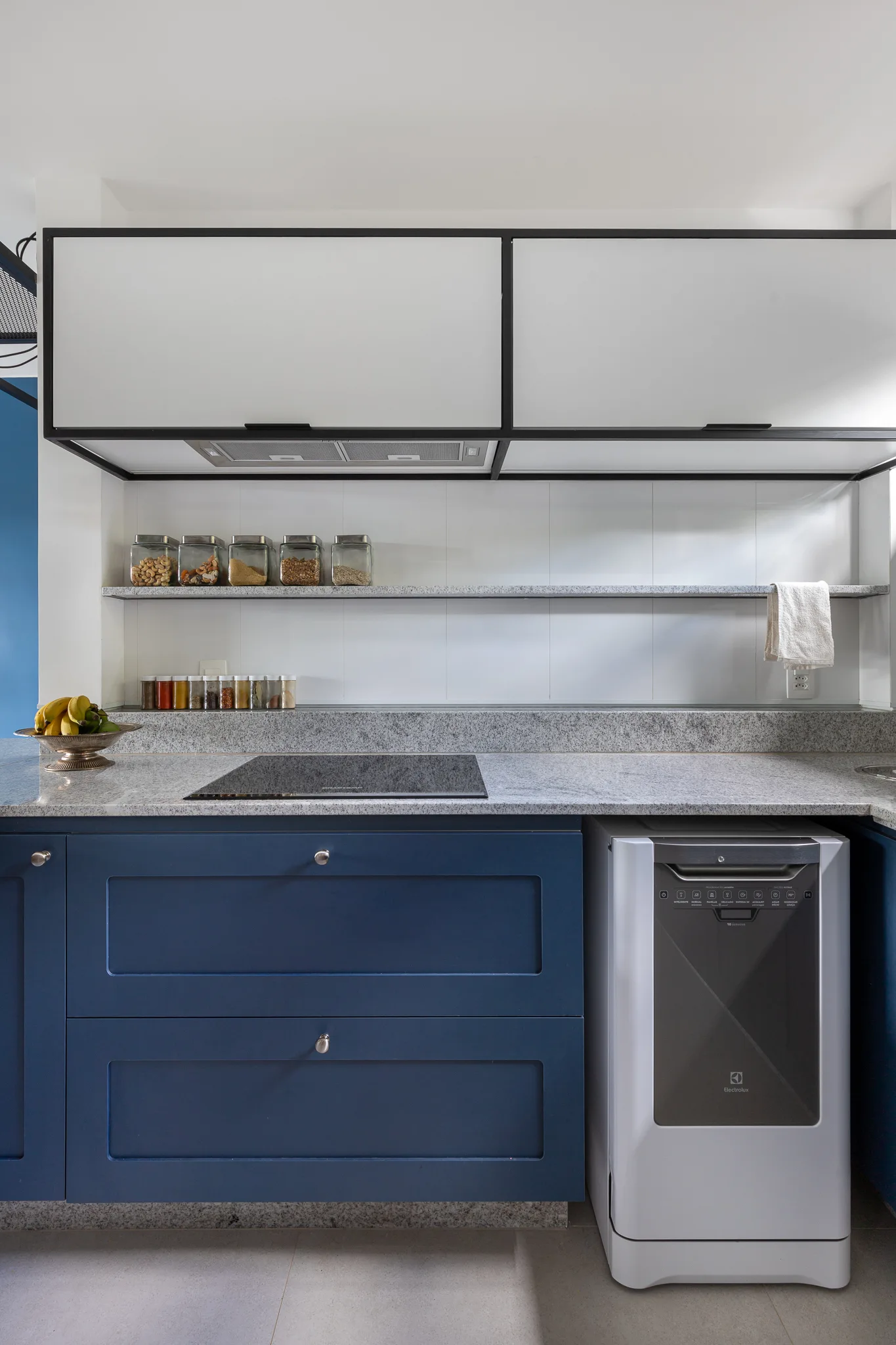 A modern kitchen features navy blue lower cabinets, speckled grey stone countertops, a matching floating shelf, and white upper cabinets with black frames.