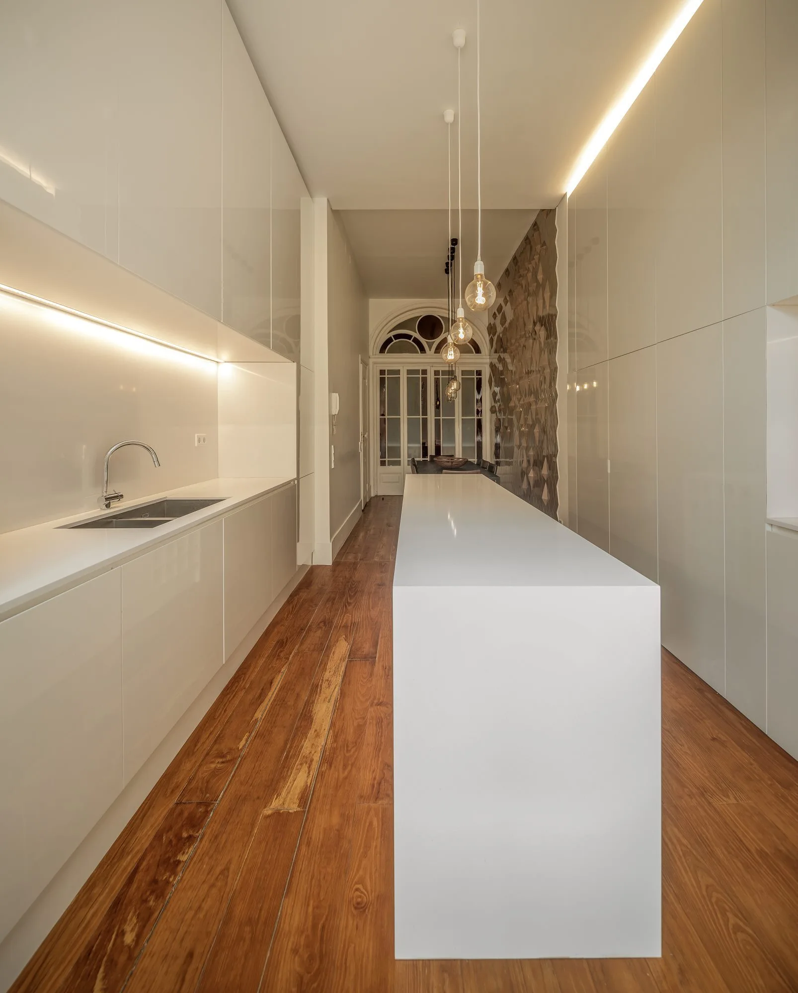 A narrow modern kitchen featuring a long white minimalist island, high-gloss cabinets with integrated strip lighting, and warm wood flooring.