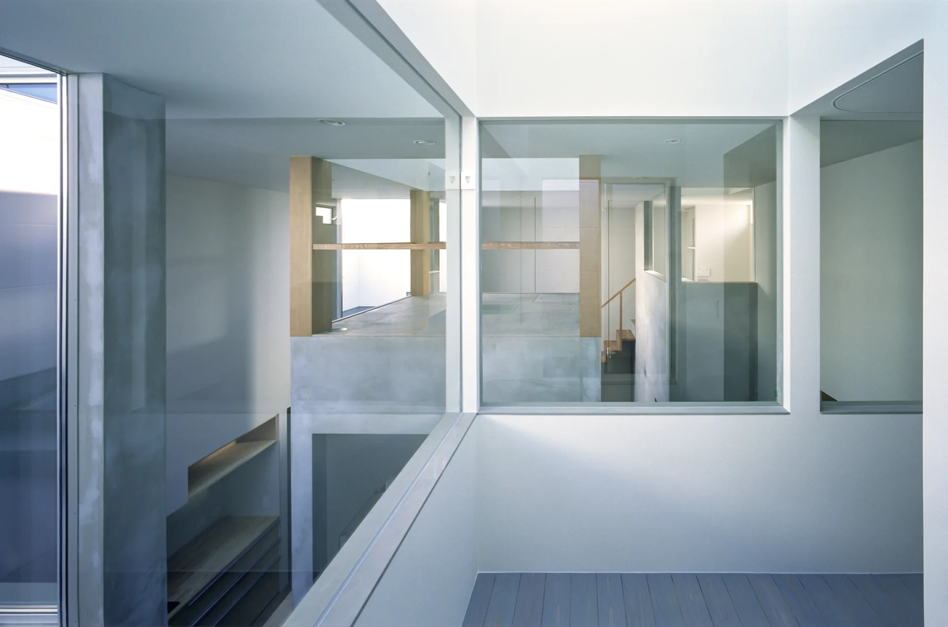 A minimalist interior featuring multi-level spaces separated by large architectural glass panels with white walls and grey plank flooring.