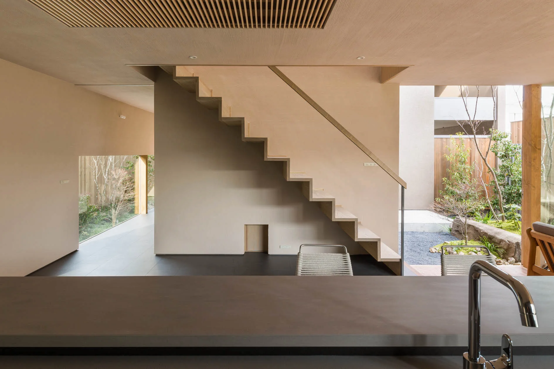 A dark gray kitchen counter in the foreground overlooks a central light wood staircase and a small outdoor courtyard with green plantings.