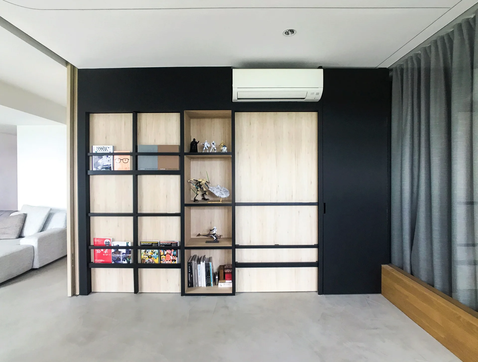 A minimalist living room features a custom black built-in wall unit with light wood shelves, displaying books and collectibles next to grey curtains.