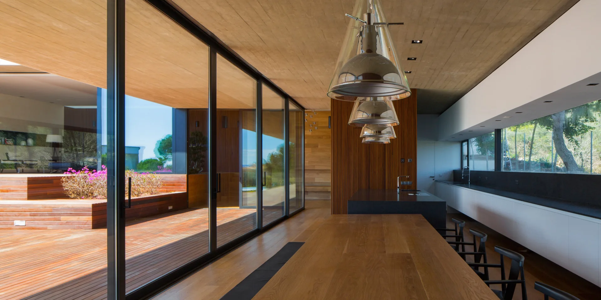 A modern kitchen featuring a long wooden dining island, white cabinets, and a wood ceiling next to sliding glass doors that open onto an outdoor deck.