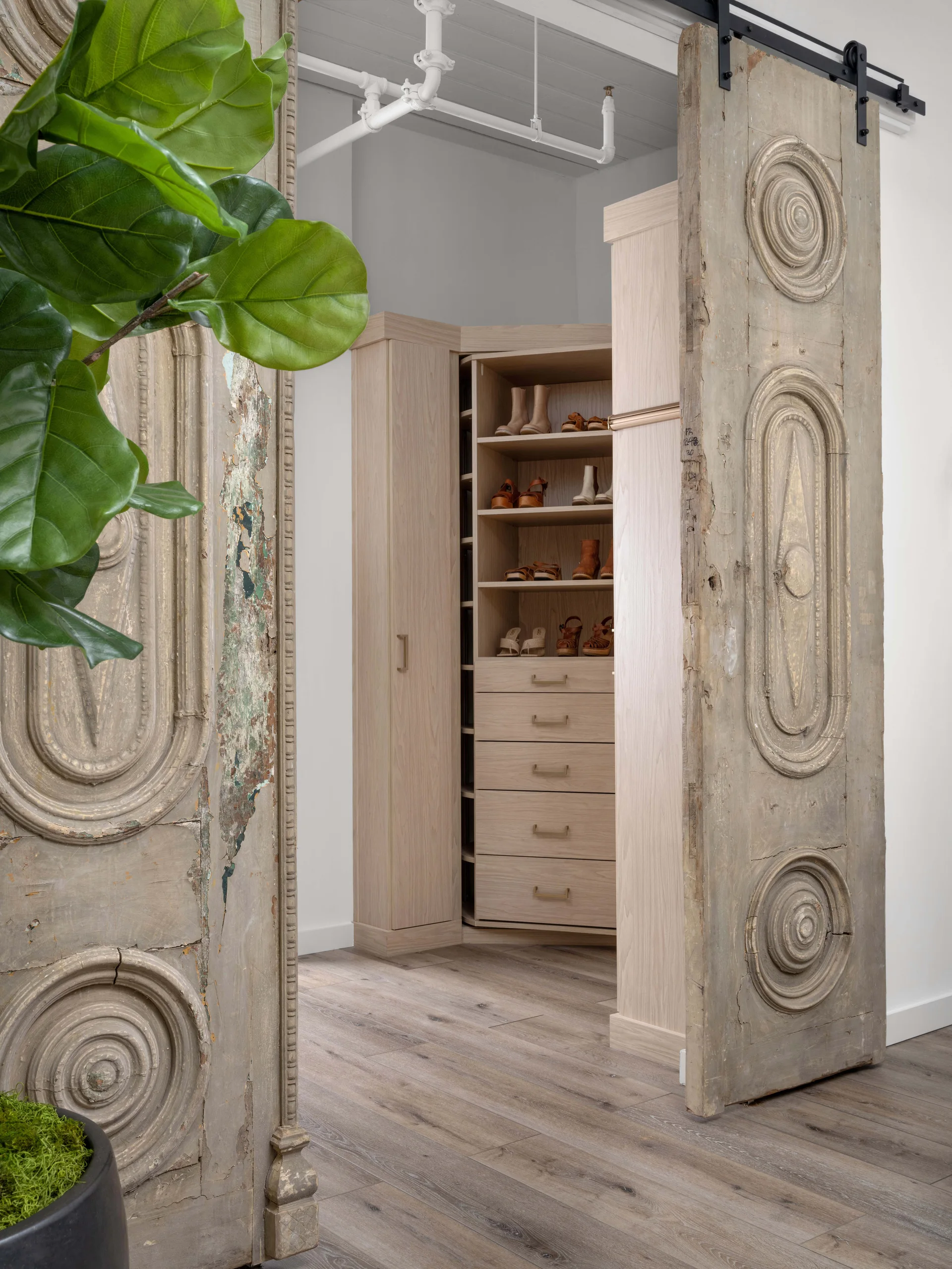 A modern walk-in closet with light wood shelving and drawers viewed through weathered antique sliding doors with carved details.