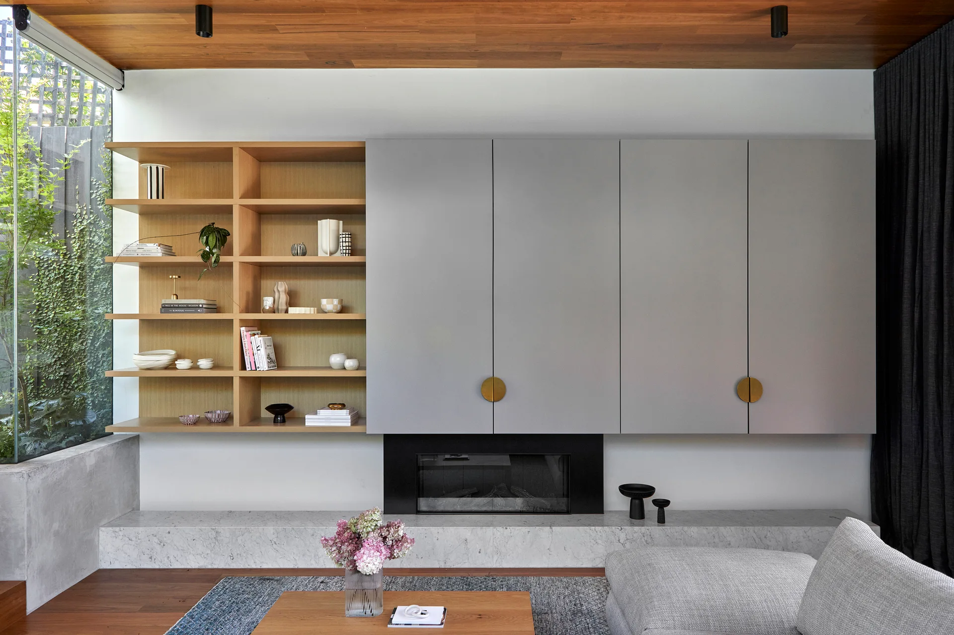 A minimalist living room features floating gray cabinets next to light wood shelves above a marble fireplace hearth and a wood-paneled ceiling.