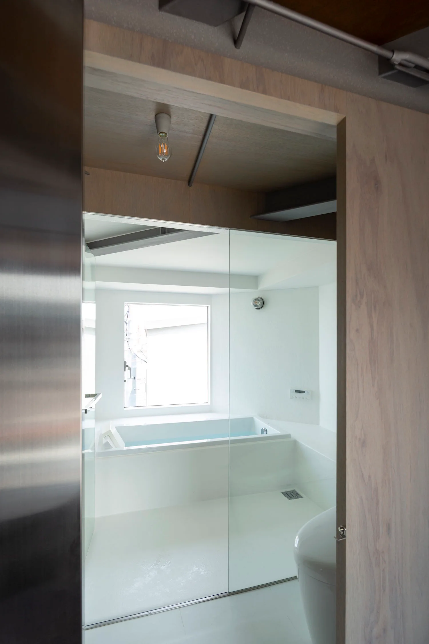 A modern white bathroom viewed through a wooden doorway, featuring a soaking tub behind a glass wall partition and a square window.
