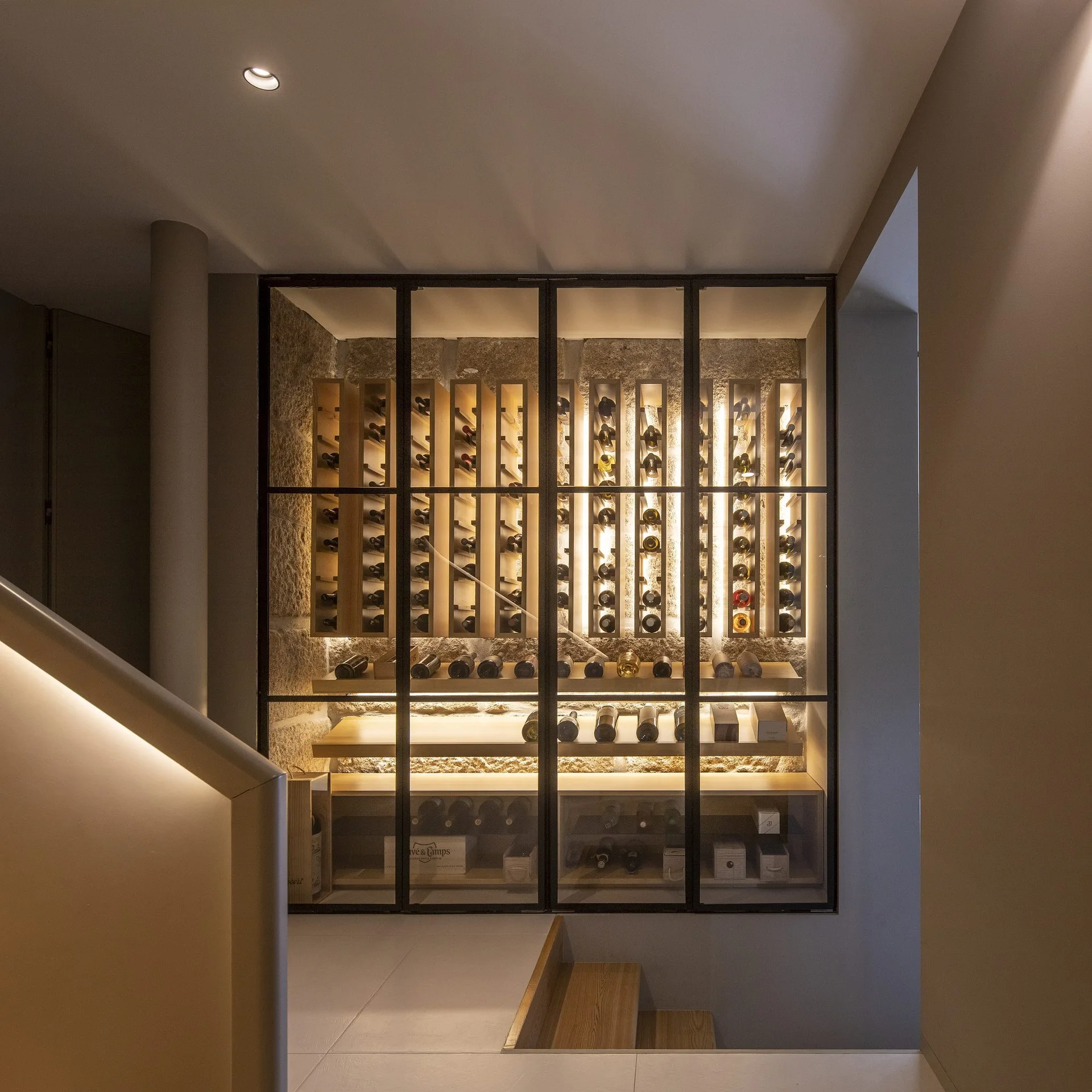 A glass-enclosed wine storage room features wooden bottle racks against a textured stone wall, situated next to a modern staircase with warm lighting.