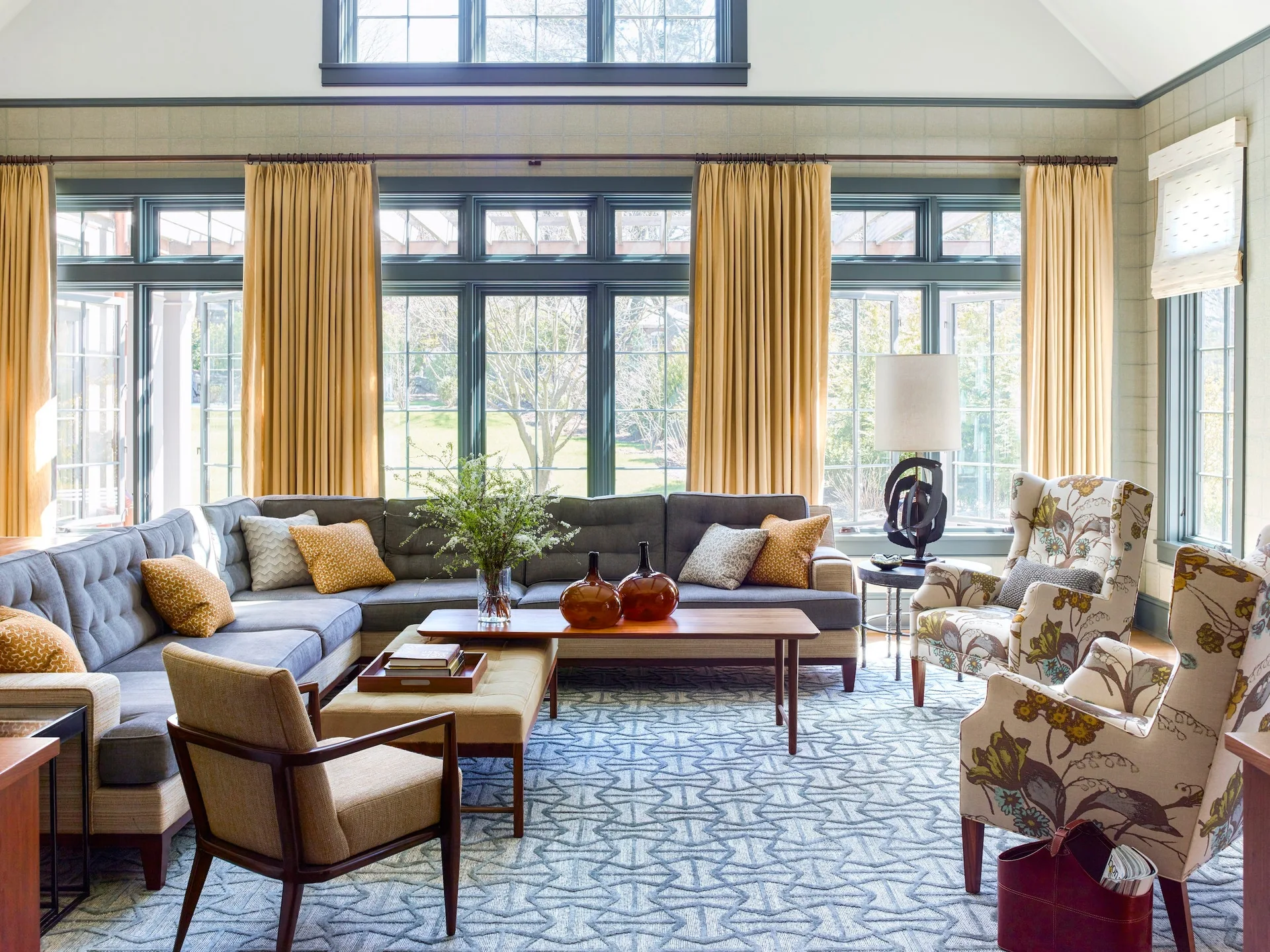 A living room with a large gray sectional sofa, gold curtains, two patterned armchairs, a wooden coffee table, and a blue geometric patterned rug.