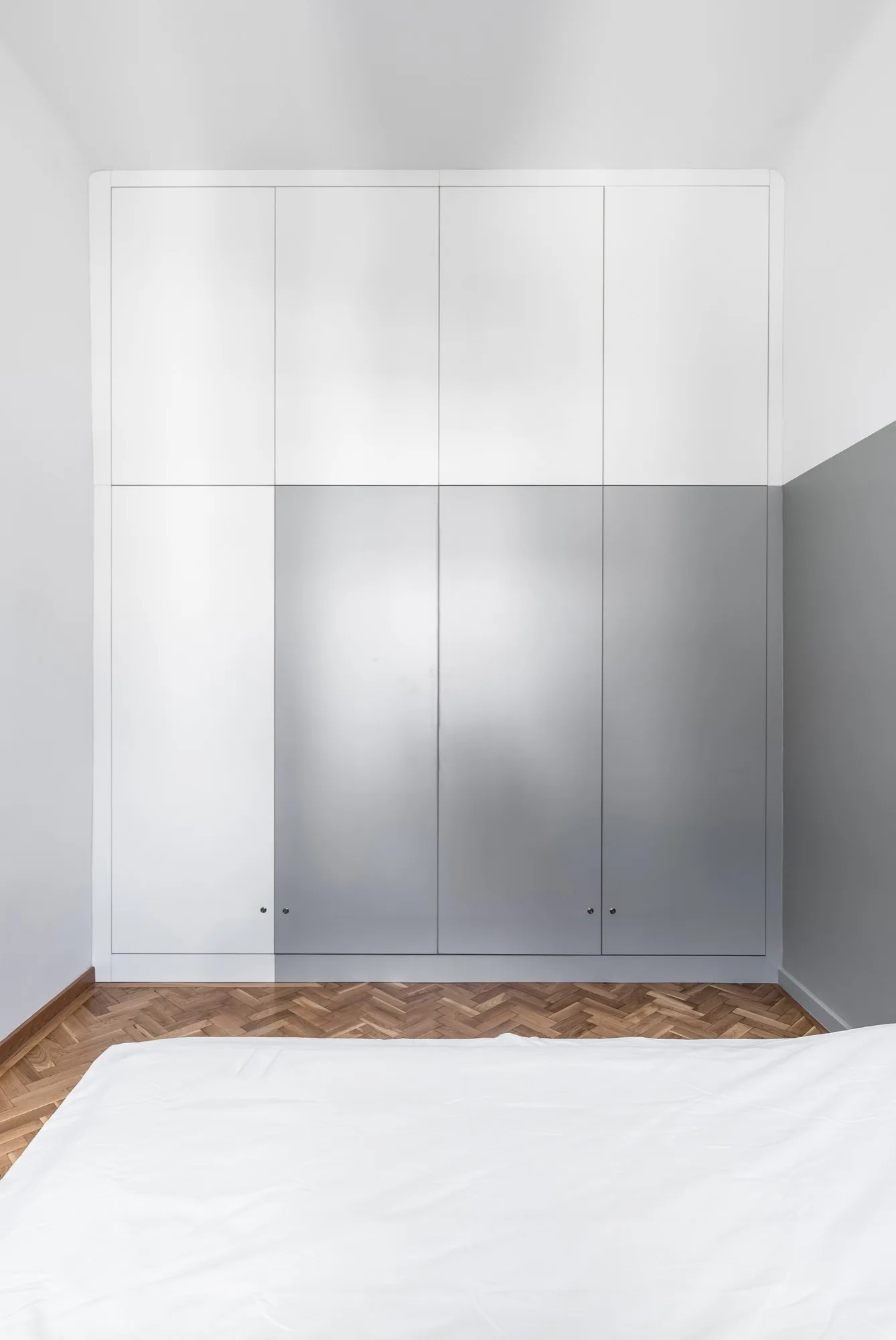 A minimalist floor-to-ceiling bedroom wardrobe features matte white and grey panels above a polished wood herringbone parquet floor.