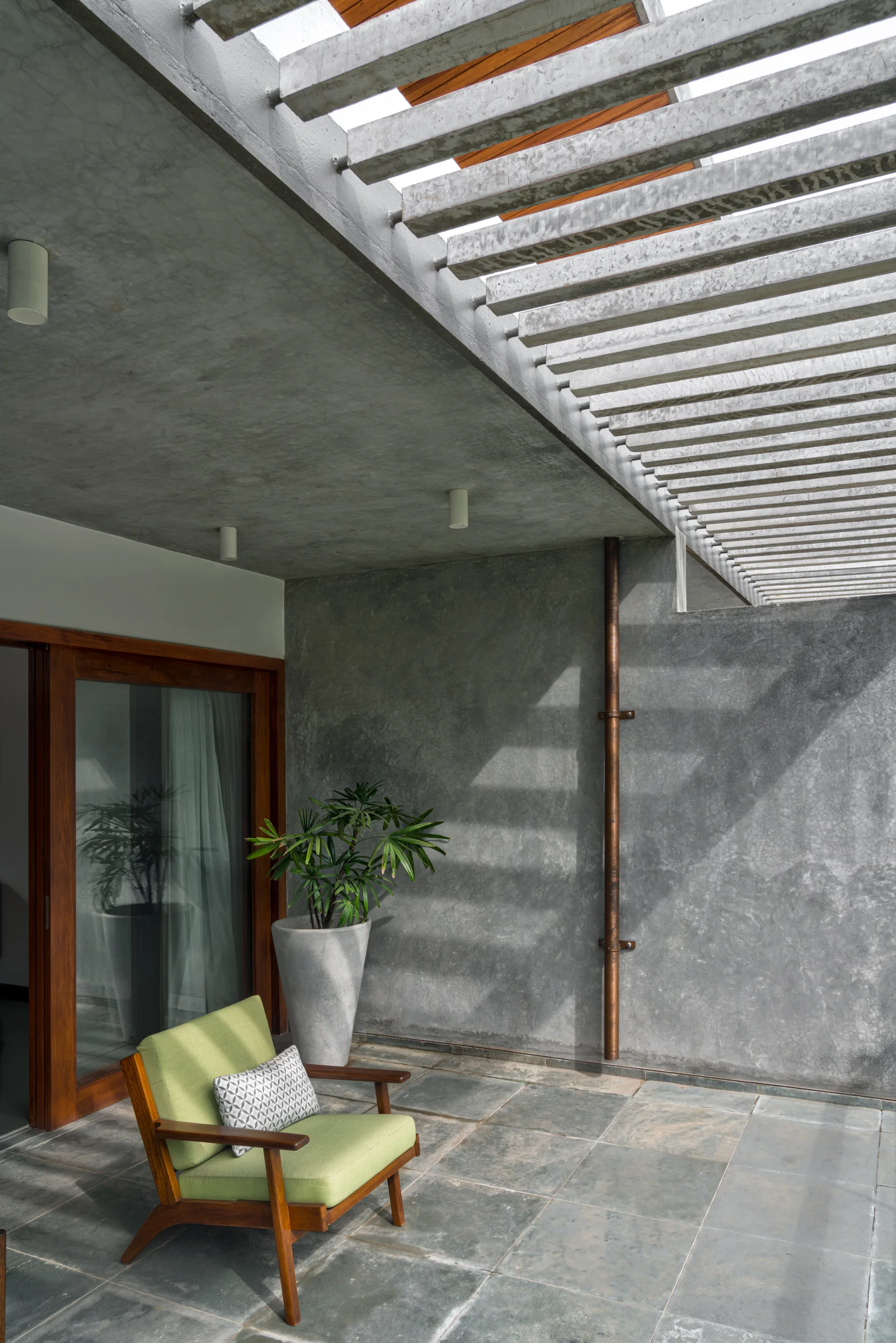 A modern concrete patio features a green armchair, a tall planter, and an exposed copper downspout under a slatted roof.