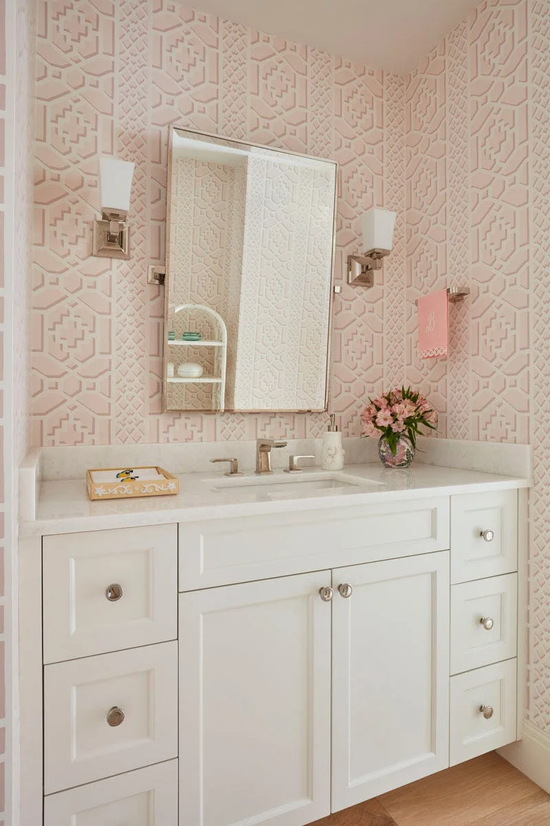 A powder room features a white shaker vanity with a stone countertop, a rectangular mirror, and blush pink geometric wallpaper.