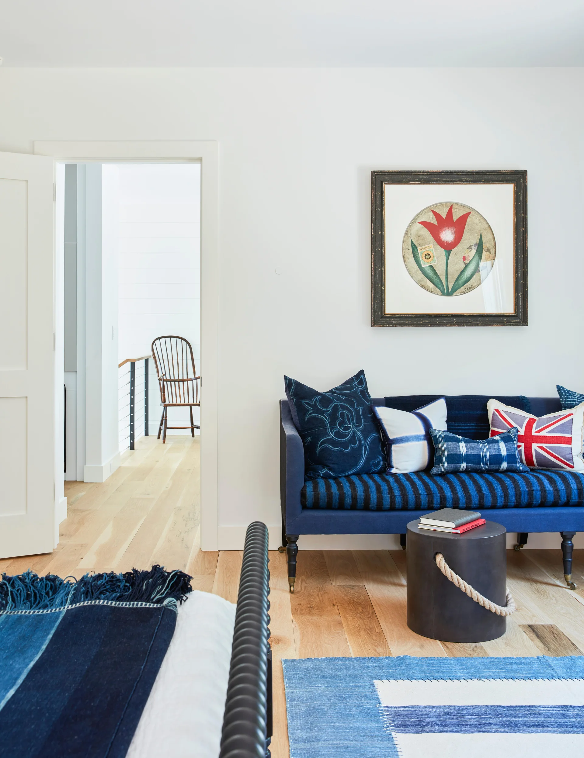 A navy blue bench with patterned pillows sits against a white wall in a bedroom next to an open doorway showing a wooden spindle chair.