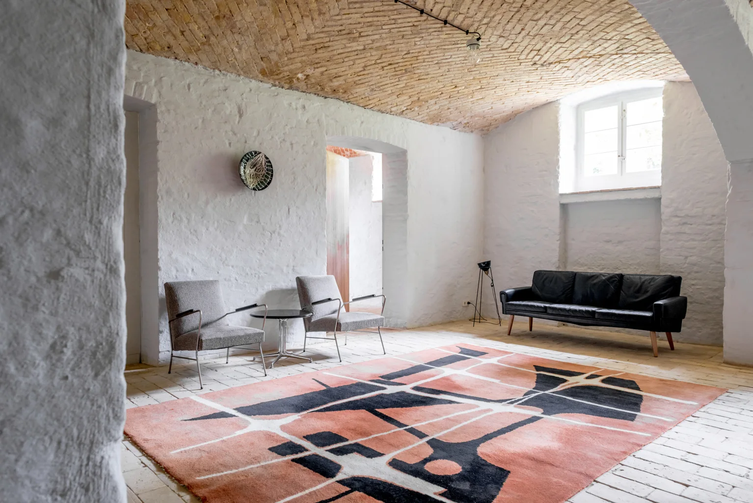 A minimalist living room features a vaulted brick ceiling, whitewashed plaster walls, a black leather sofa, and a large peach and black geometric rug.