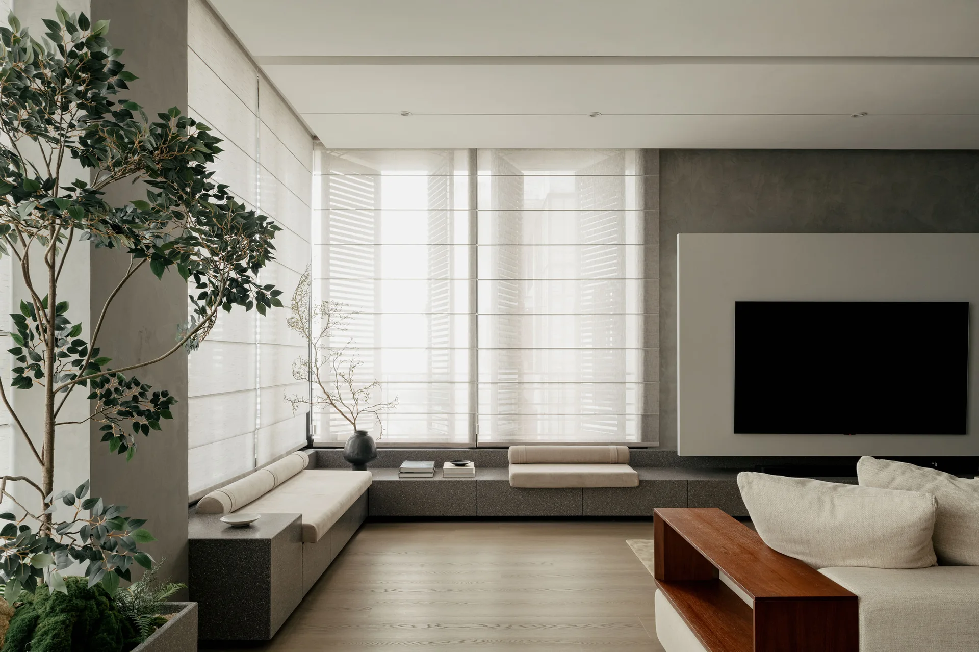 A minimalist living room features a low stone bench beneath windows with light fabric shades, next to a neutral-toned sofa and wall-mounted television.