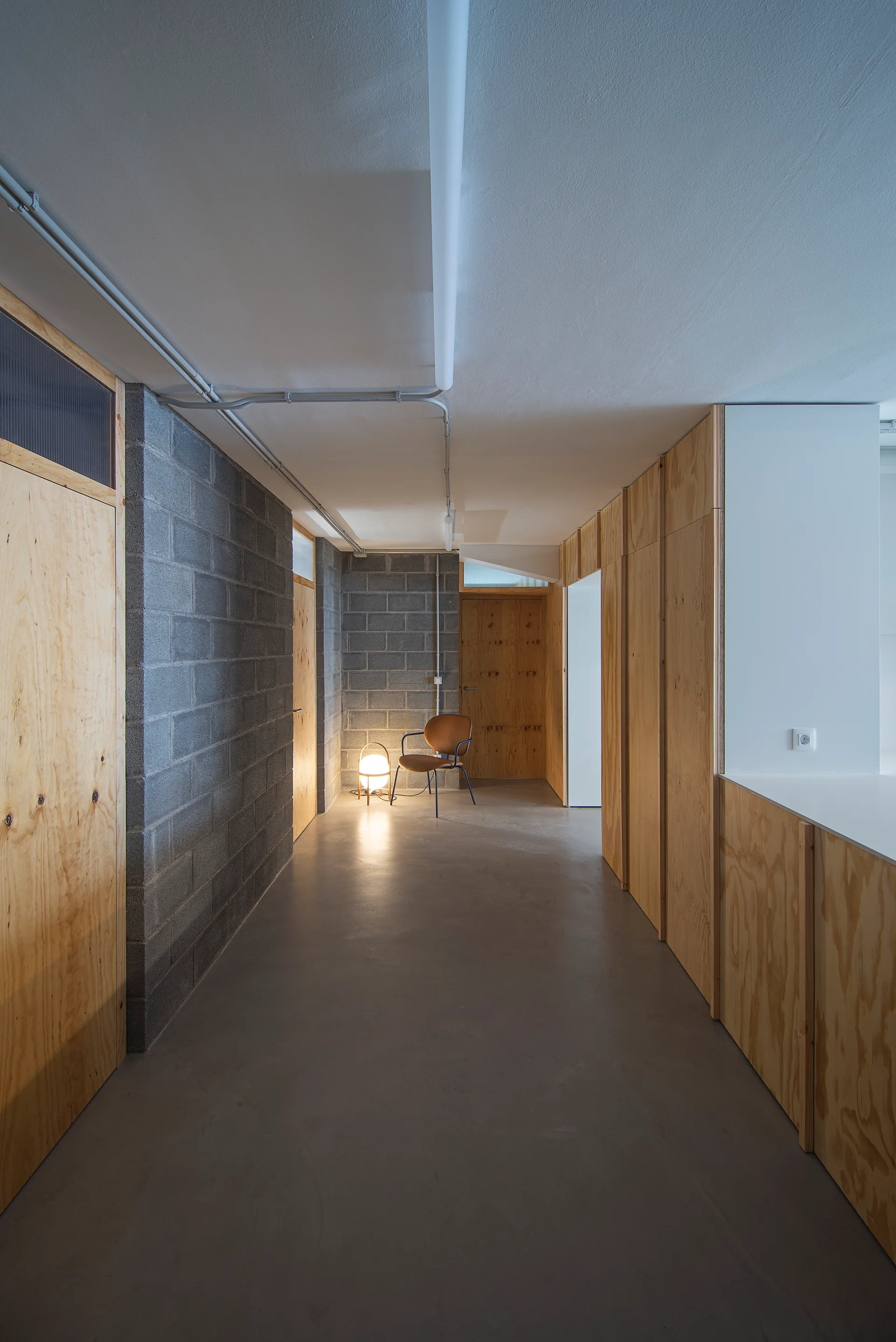 A long hallway featuring a concrete block wall on the left, natural plywood paneling on the right, and a glowing floor lamp at the far end.