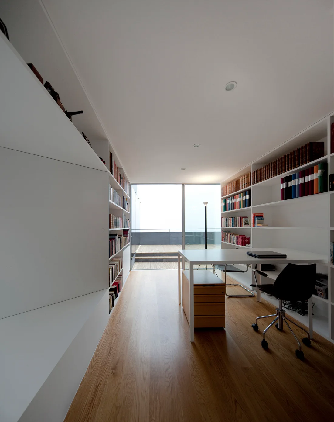 A minimalist home office features symmetrical white built-in bookshelves, a white desk with a black chair, and glass doors leading to a wooden deck.
