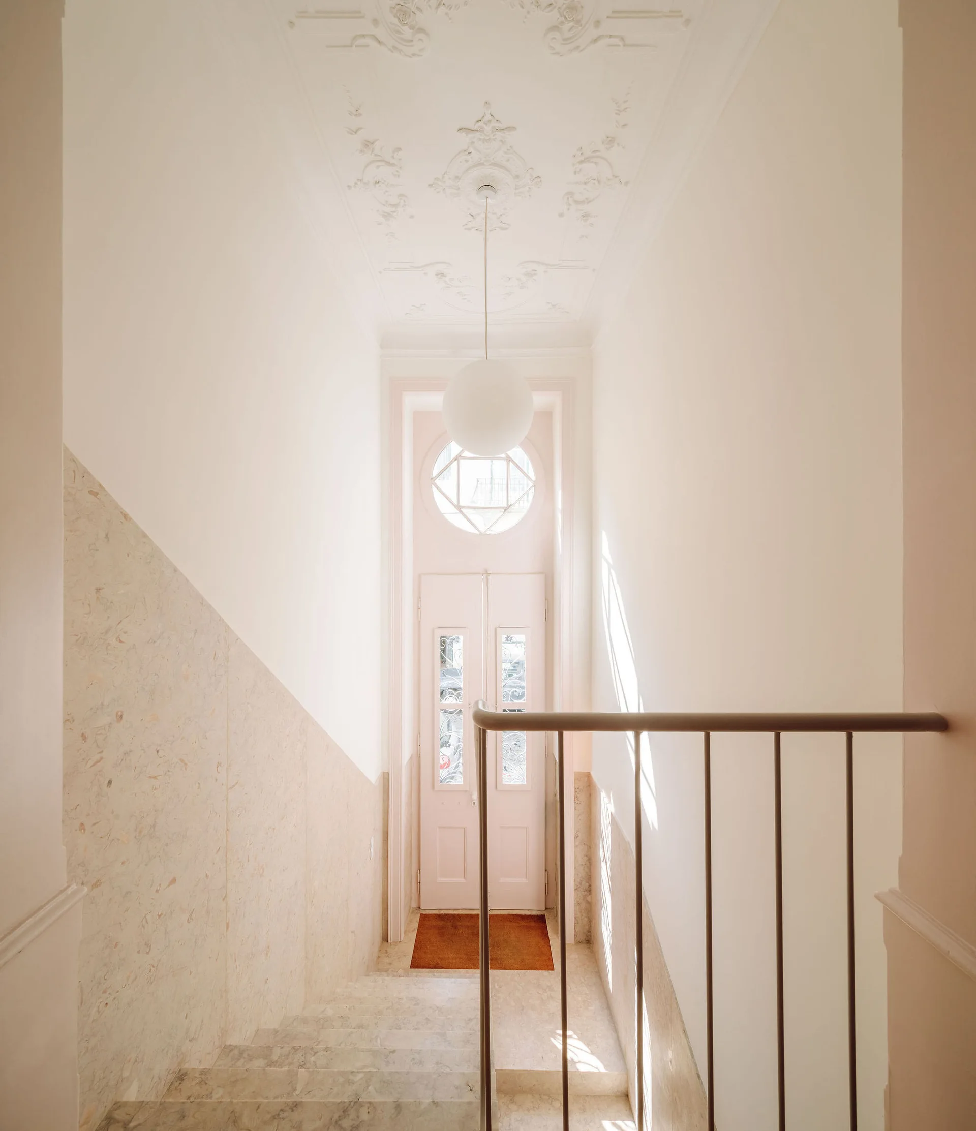 A light-filled stairwell with stone wall paneling, a minimal metal railing, an ornate plaster ceiling, and a hanging white globe pendant.