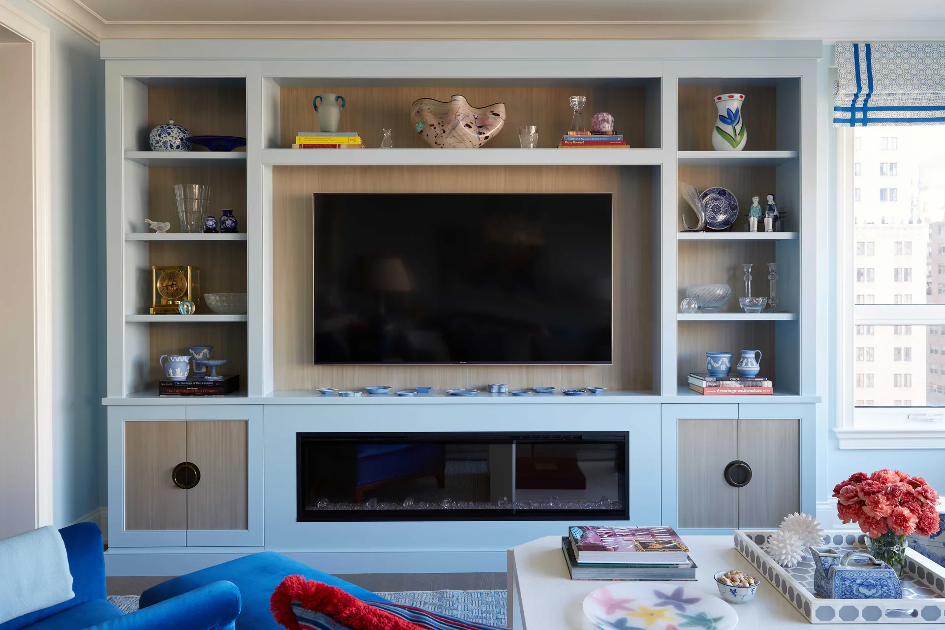 Light blue custom built-in shelving surrounds a wall-mounted television and electric fireplace in a bright living room decorated with colorful vases.