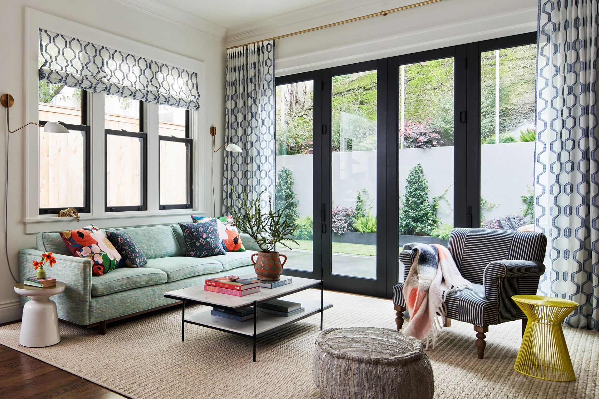 A bright living room features a green-blue sofa, a striped armchair, and geometric print curtains next to large black French doors opening to a garden.