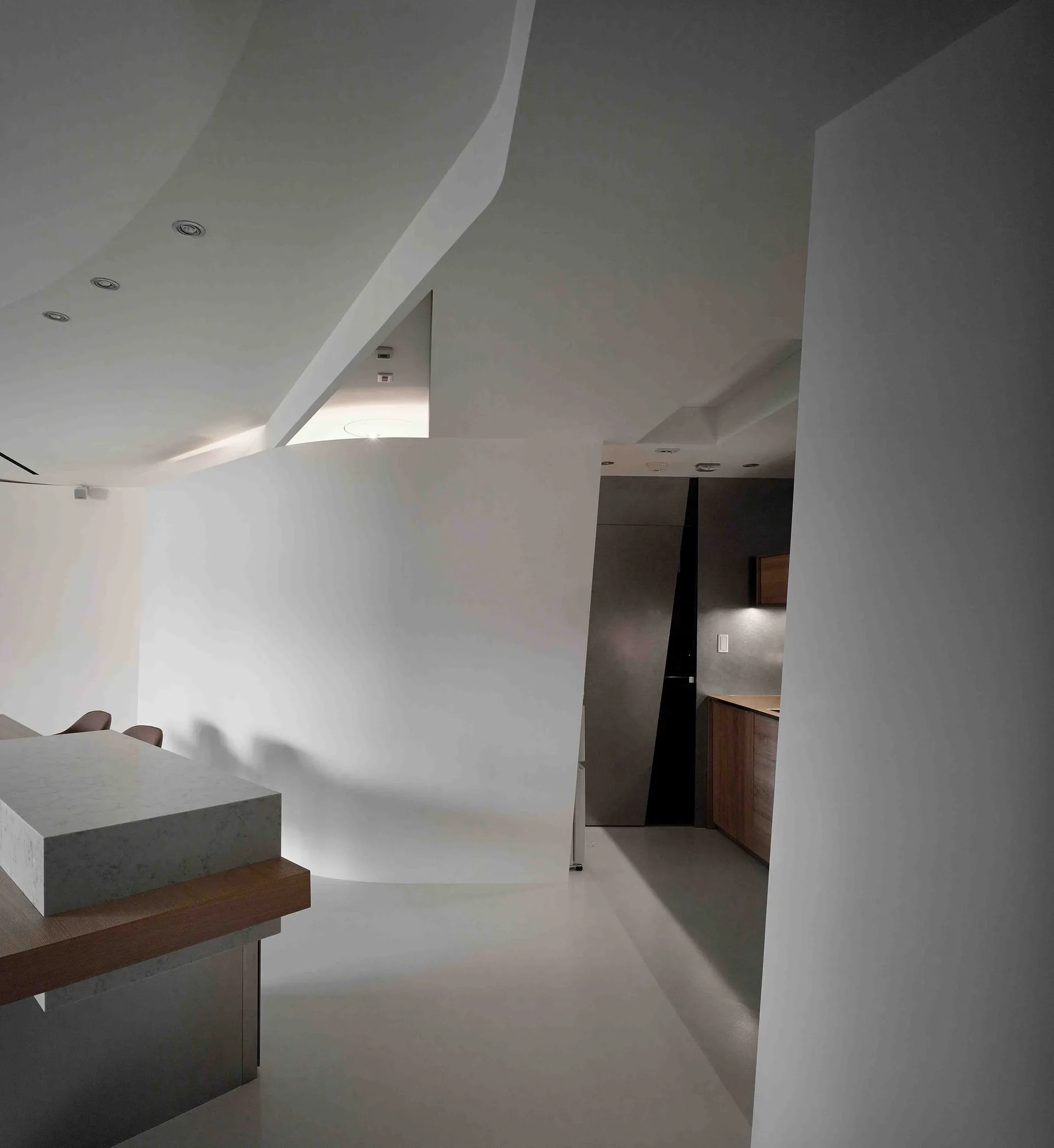 A minimalist kitchen features a stone-topped island, wood-toned cabinets, a large white curved wall, and an angled sculptural ceiling.