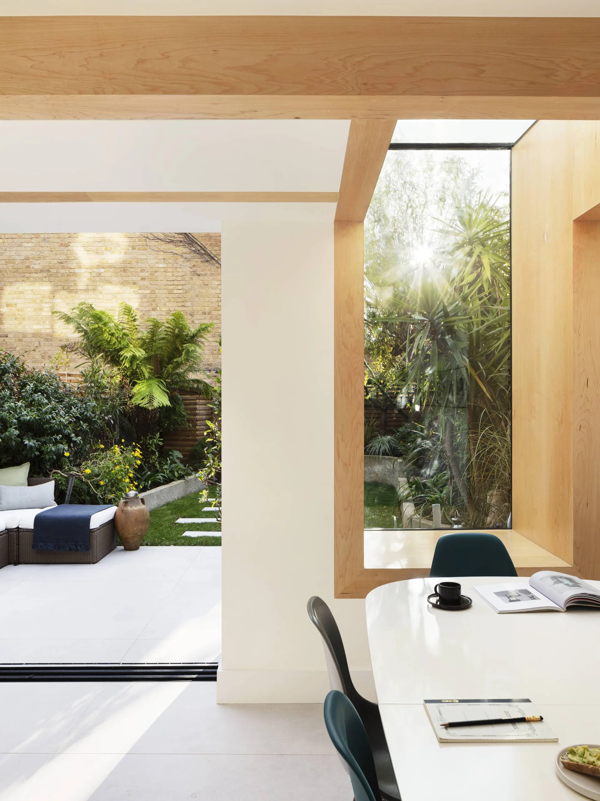 A modern dining room features a white table, dark chairs, and exposed ceiling beams next to a large wood-trimmed window looking out onto a garden.